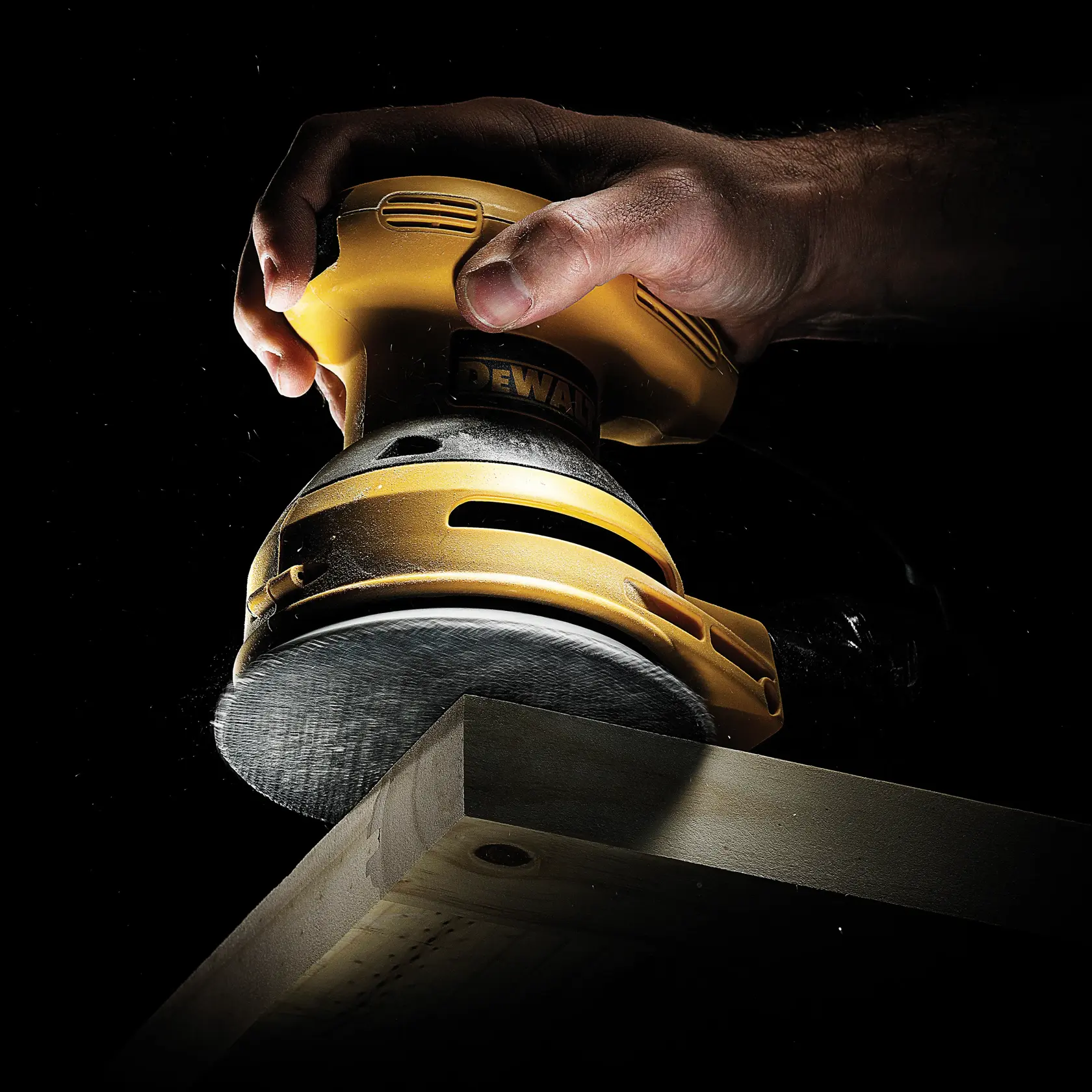 Hand holds DEWALT sander polishing wooden edge against dark background.
