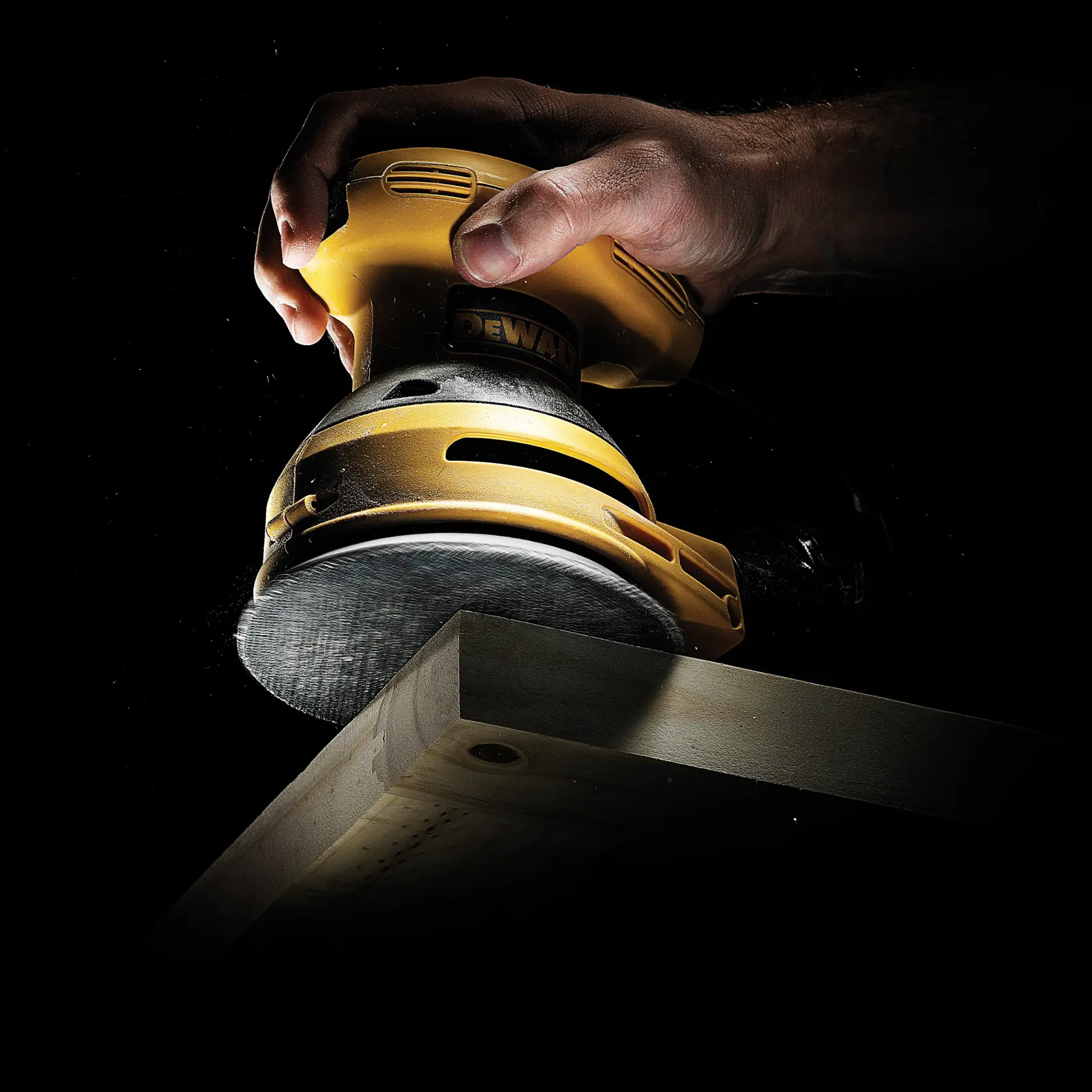 Hand holding a DEWALT sander, smoothing the edge of a wooden plank against a dark background.