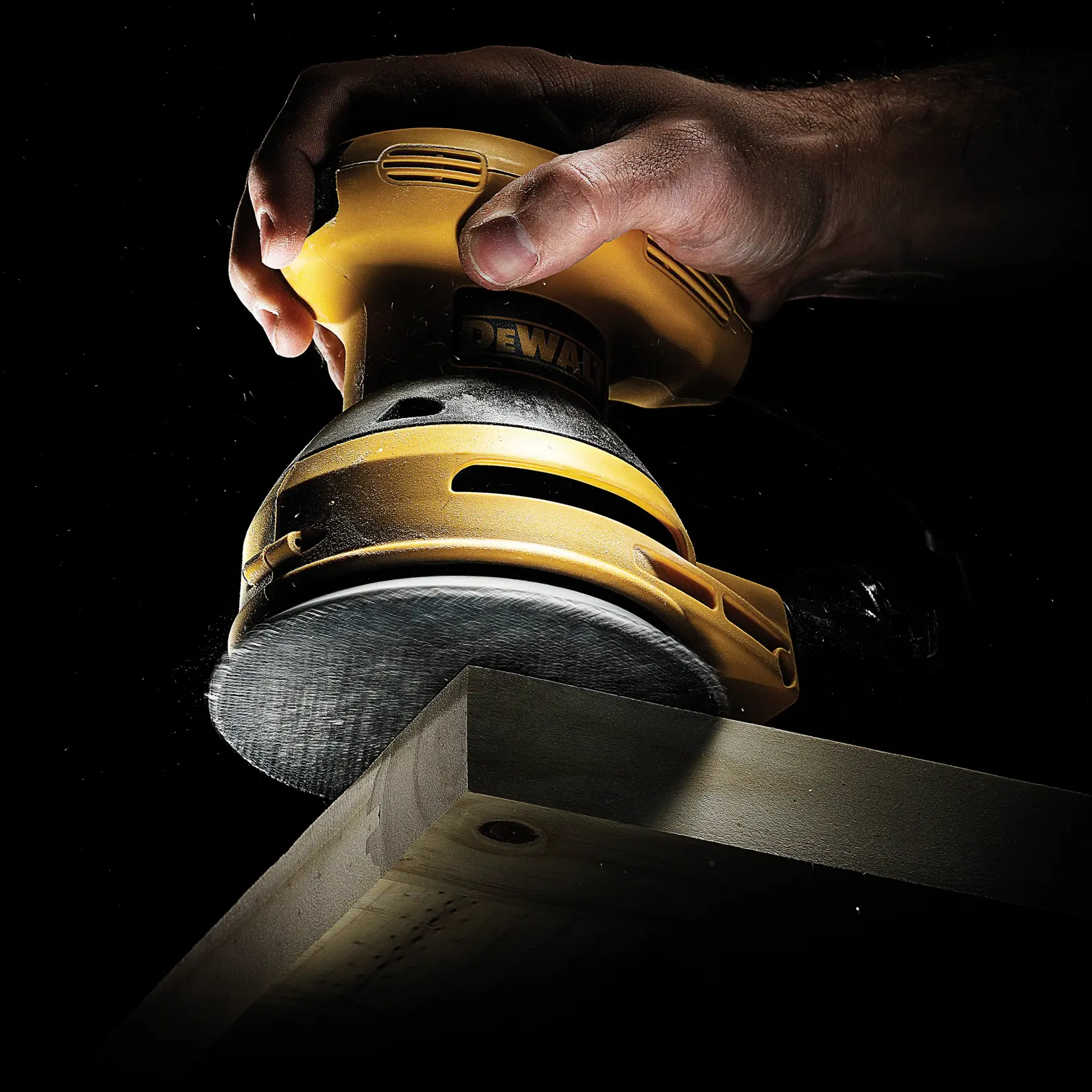 Hand holding a DEWALT sander, smoothing the edge of a wooden plank against a dark background.