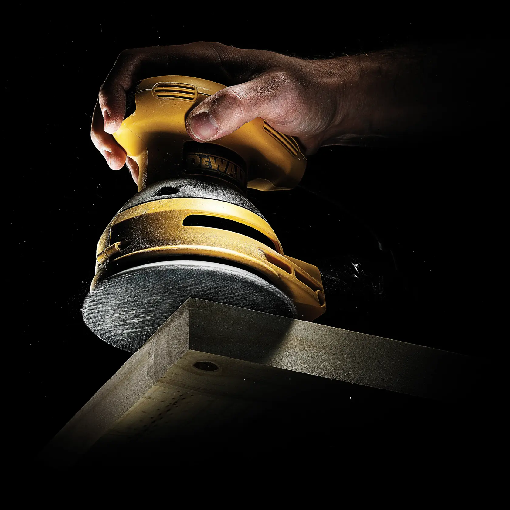 A hand uses a DEWALT orbital sander to smooth the edge of a wooden board against a dark background.