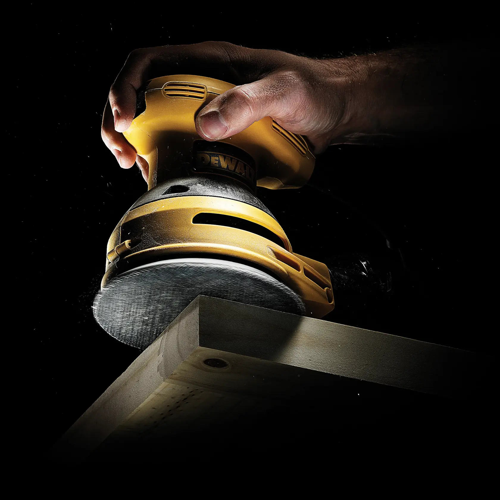 Hand using DEWALT sander to smooth the edge of a wooden board against a black background.