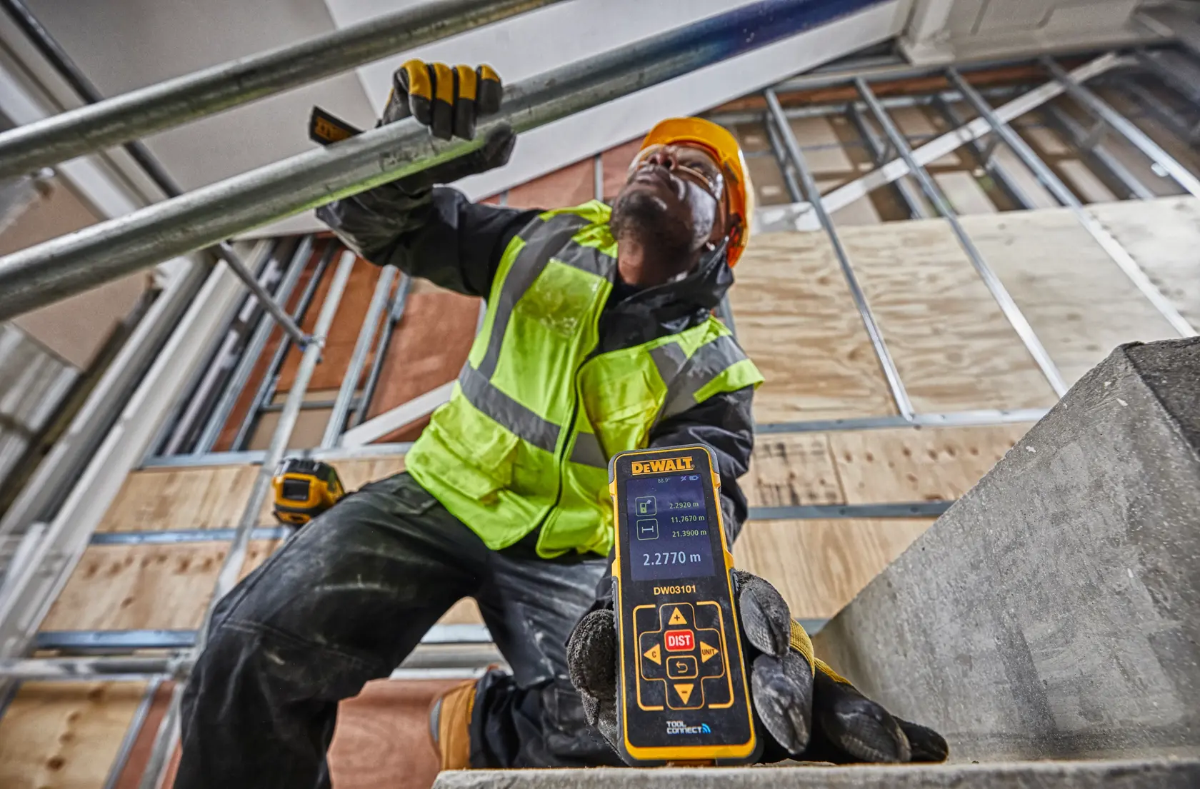 A construction worker wearing a yellow safety vest, helmet, and gloves is holding a DEWALT DW03101 laser distance measurer on a building site. The focus is on the device which displays a measurement on its screen.