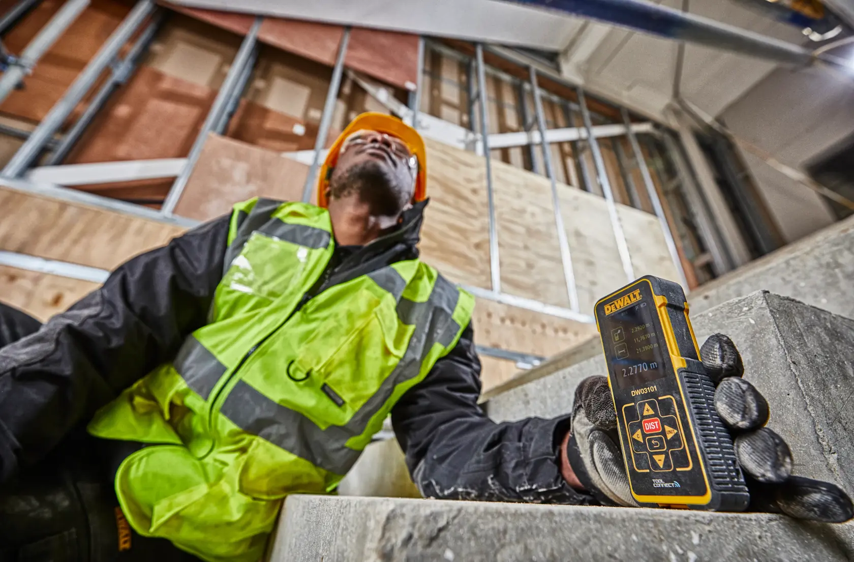 Worker holds DEWALT DW03101 laser distance measurer at a construction site with unfinished walls.