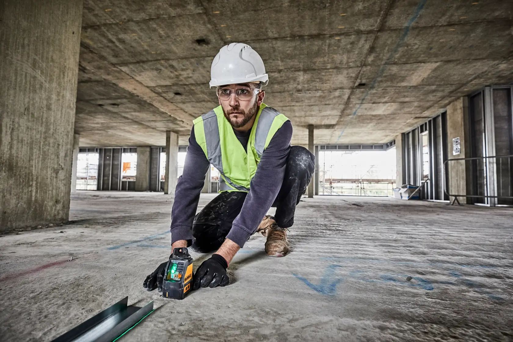 A person wearing a safety helmet, high-visibility vest, and gloves is kneeling on the floor of a large construction site