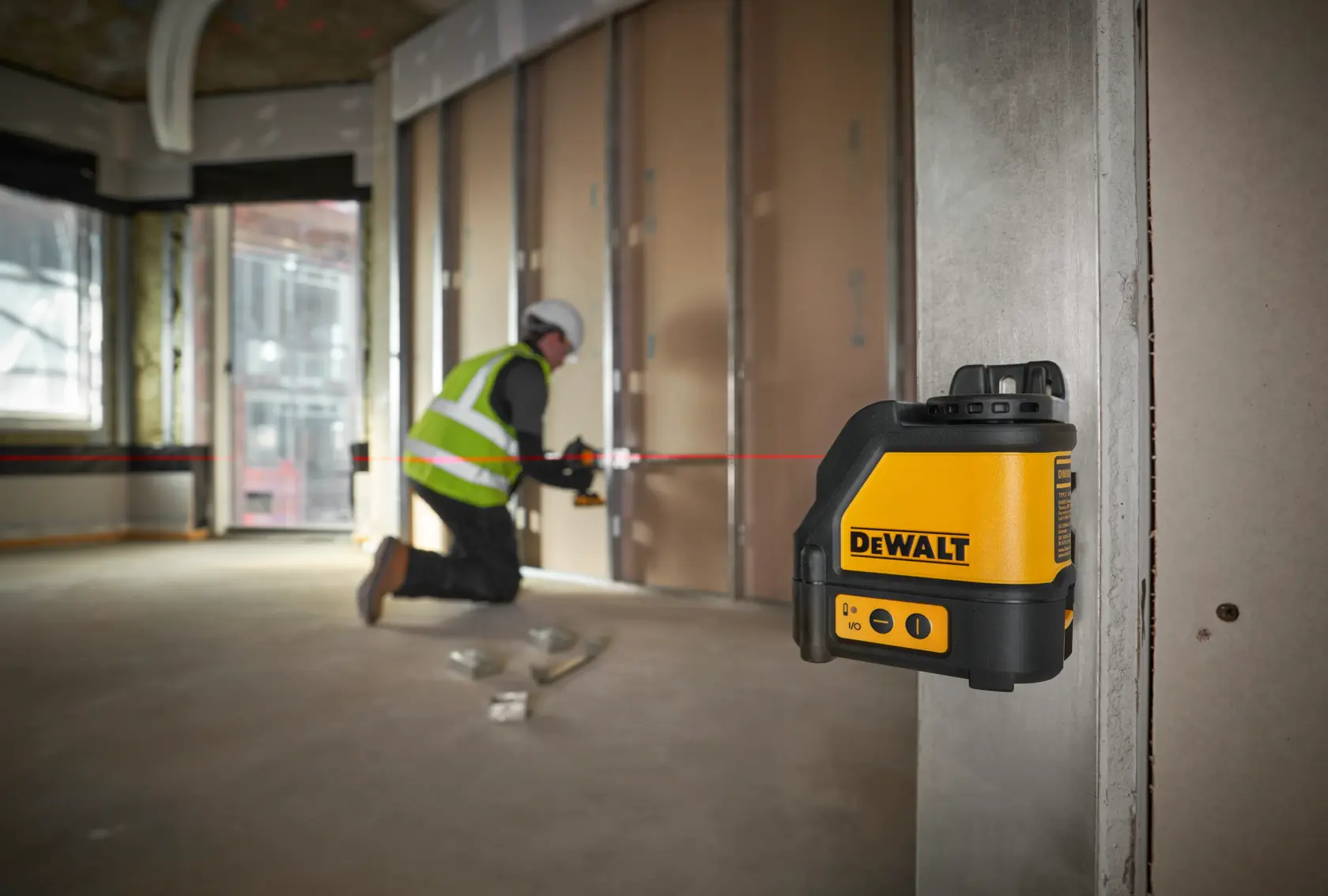 DEWALT DW088K laser level projects a red line in a construction area with a worker installing drywall.