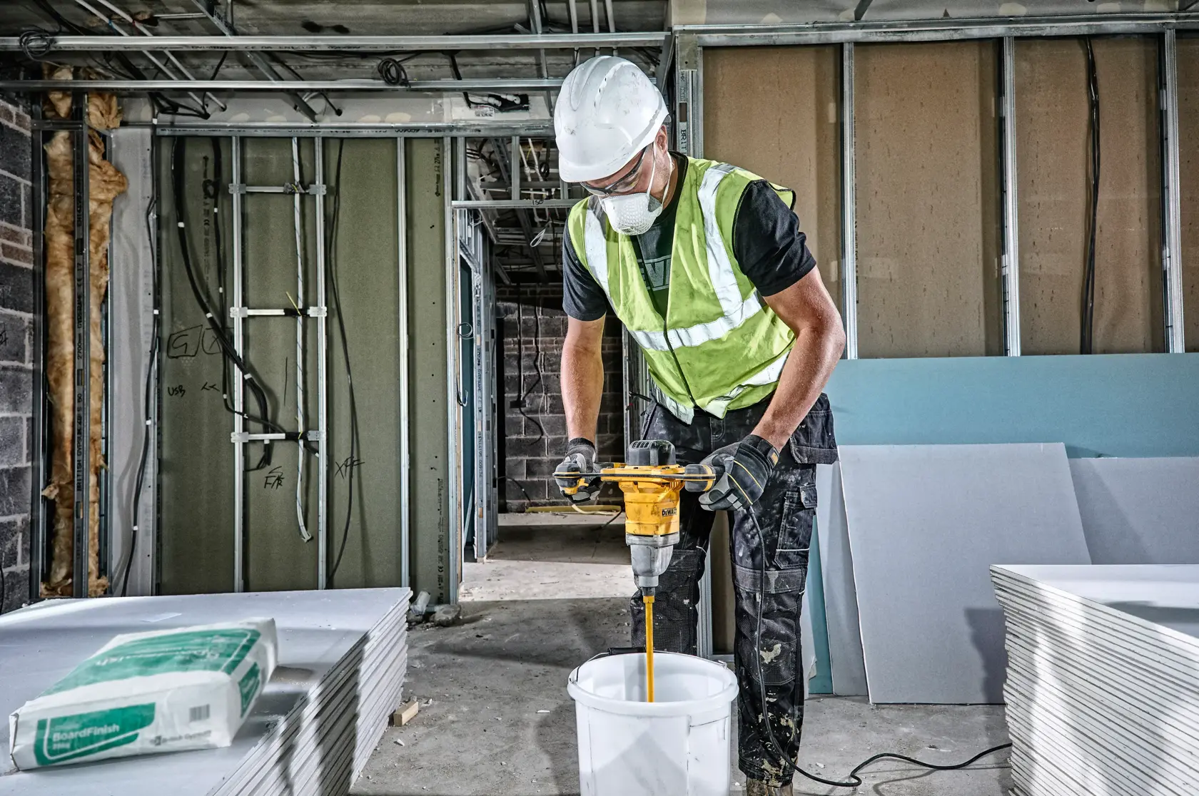 Worker uses a DEWALT DWD241 drill to mix materials in a bucket on a construction site.