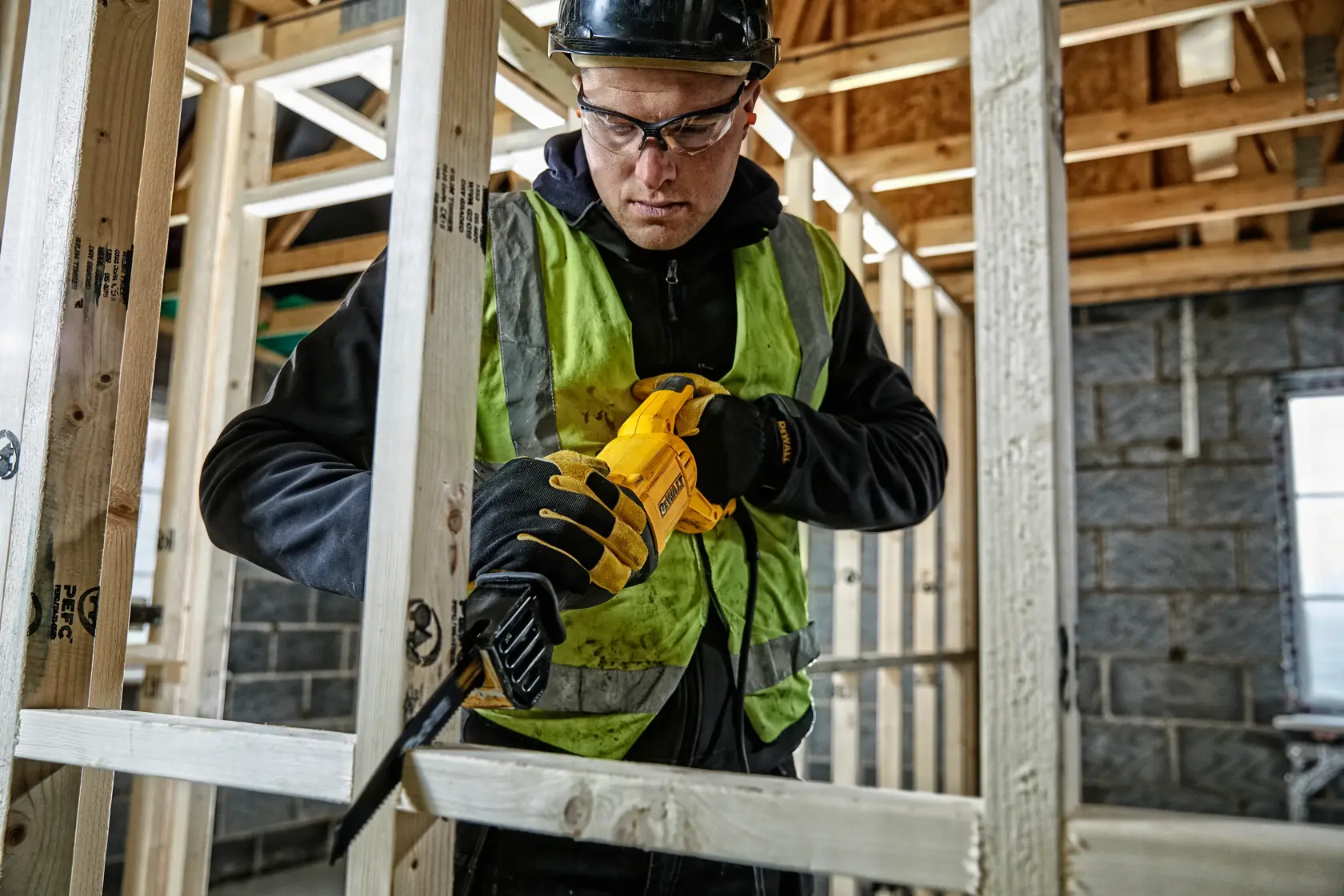 Worker uses a yellow DEWALT reciprocating saw to cut wood frame at a construction site, wearing safety gear.