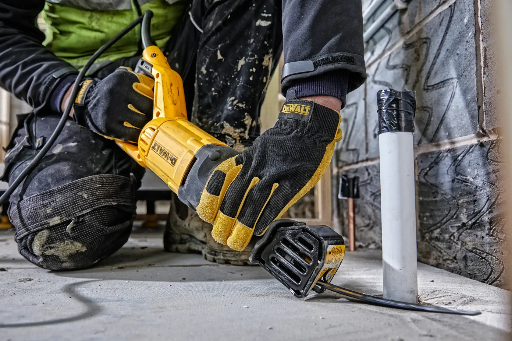 Person uses DEWALT reciprocating saw to cut pipe on construction site, wearing gloves and kneepads.