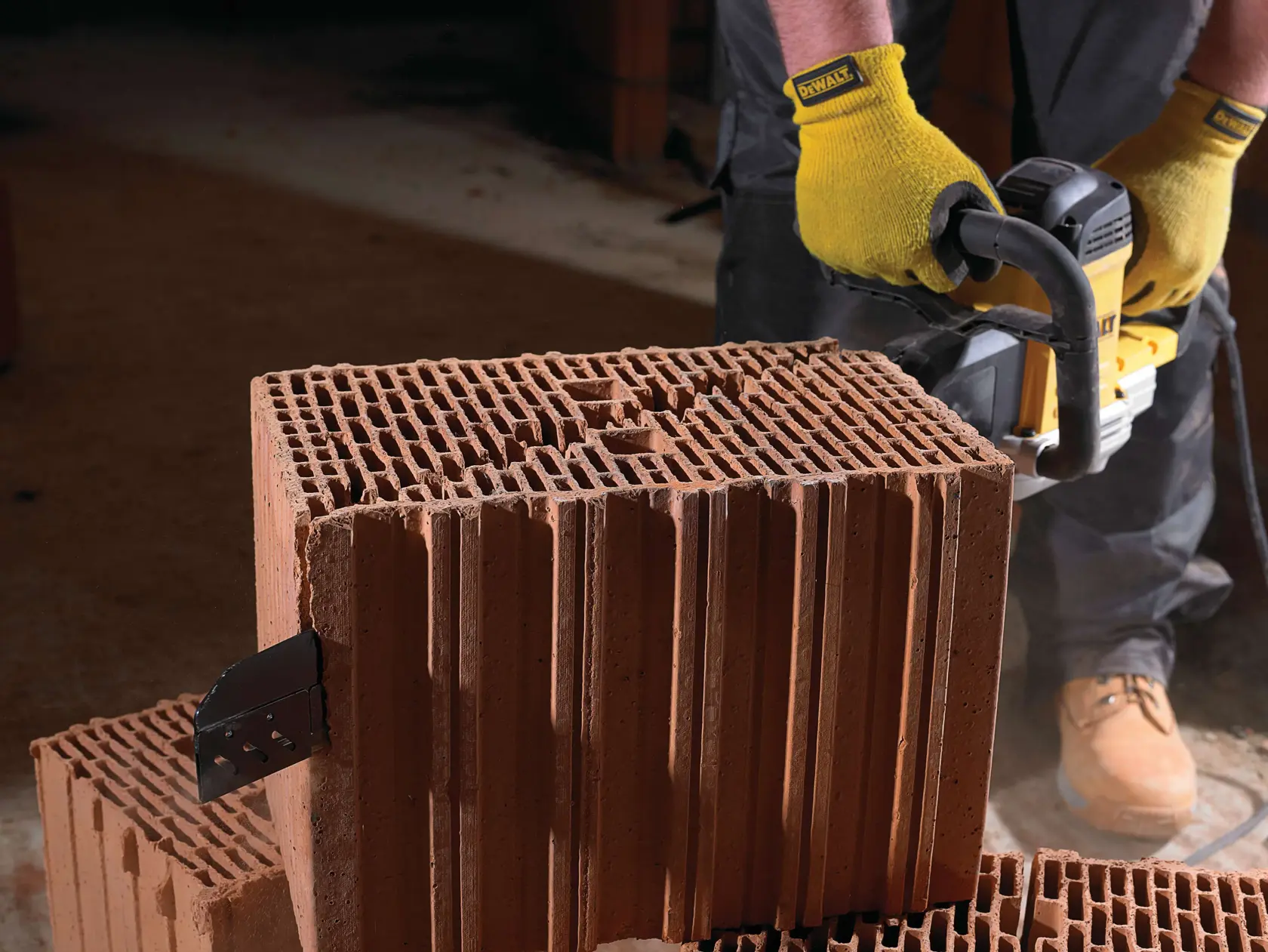 Person cutting a red brick block with a DEWALT power saw, wearing yellow gloves and work boots.