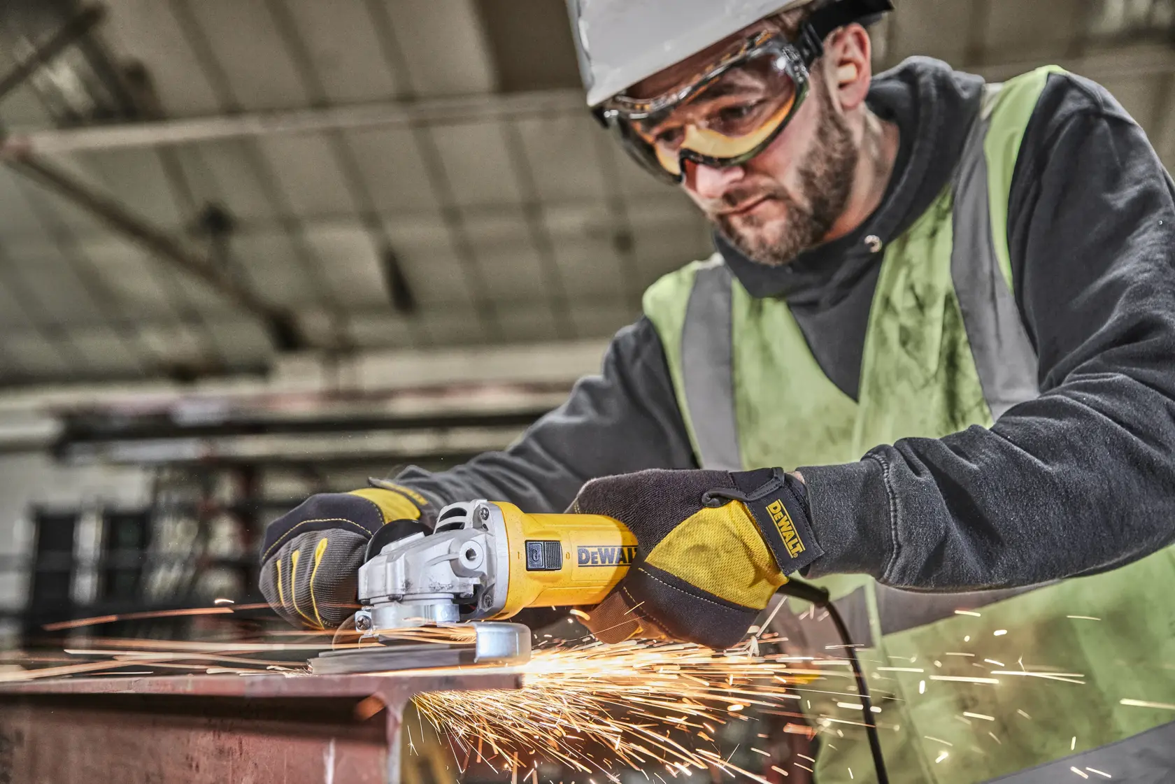 Worker grinds metal with DEWALT DWE4036 angle grinder, creating sparks in an industrial workshop.