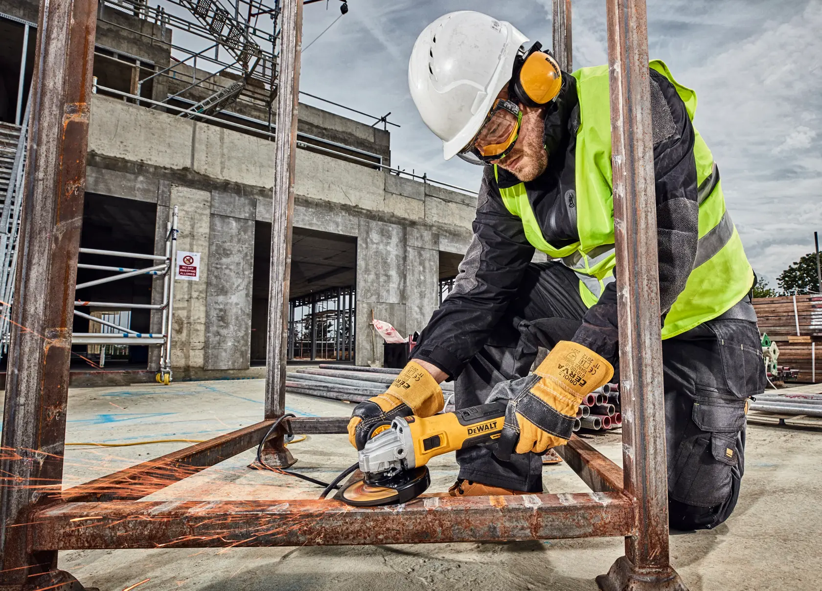 Worker uses DEWALT DWE4357 grinder on metal frame at construction site, wearing safety gear.