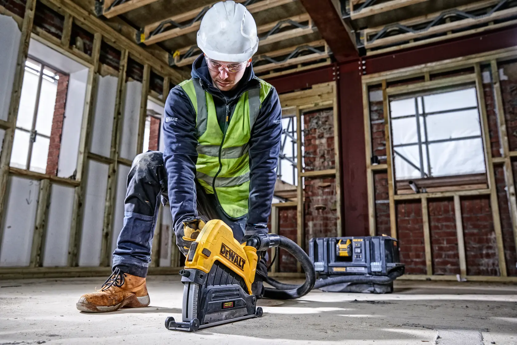 Operaio usa utensile DEWALT a terra in cantiere, con protezioni. Cassetta degli attrezzi sullo sfondo.