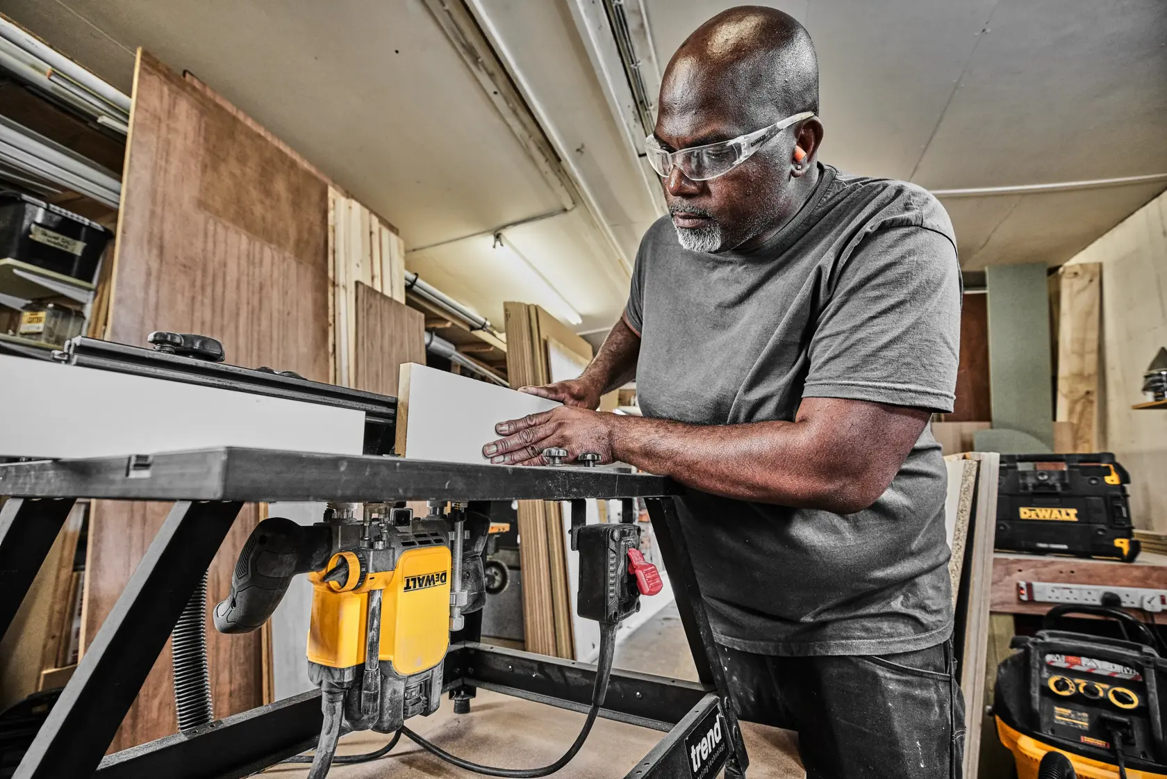 Person using a DEWALT DWE627KT router on a workshop table for woodworking, with toolboxes in the background.