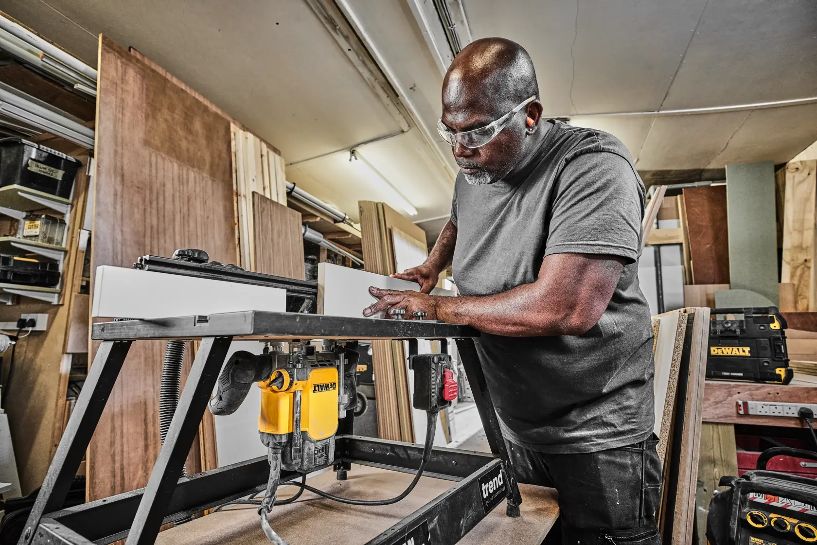 A person uses a DEWALT router tool in a workshop, working with wood on a sturdy table.