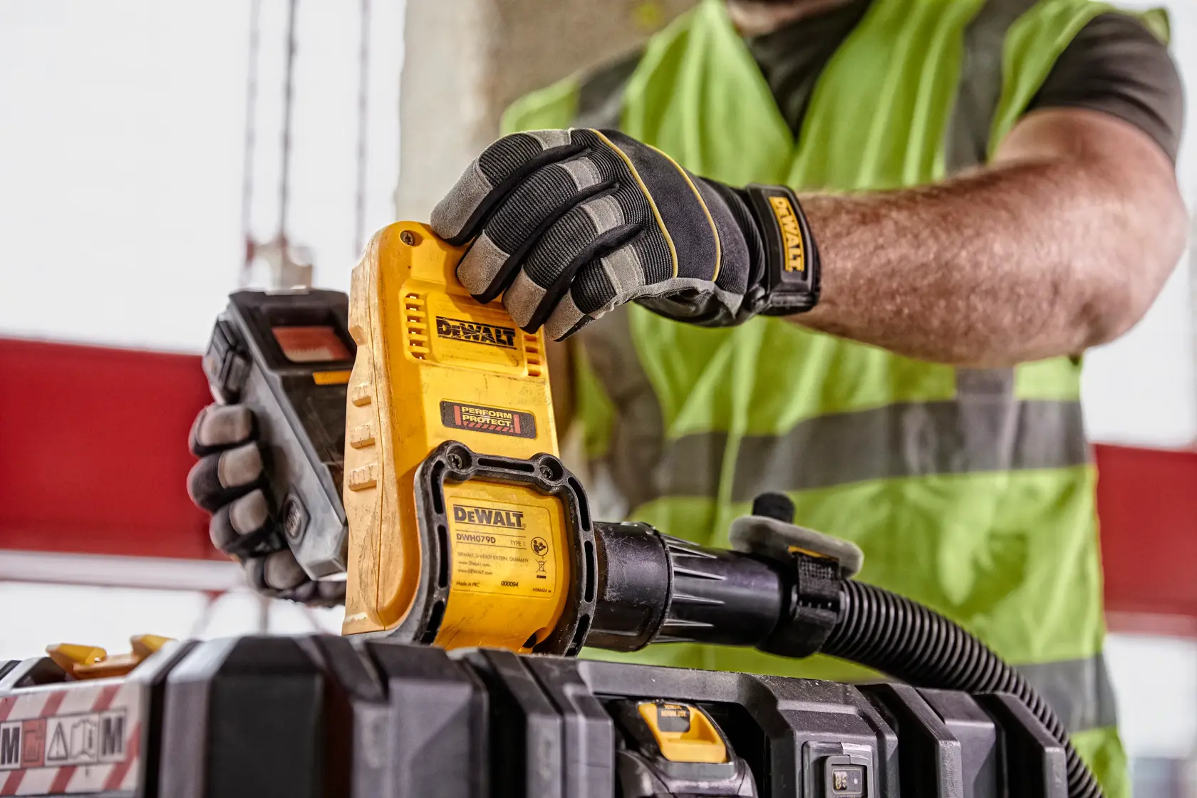 Person wearing gloves operates DEWALT DWH079D tool near a toolbox, hose attached. High-visibility vest visible.