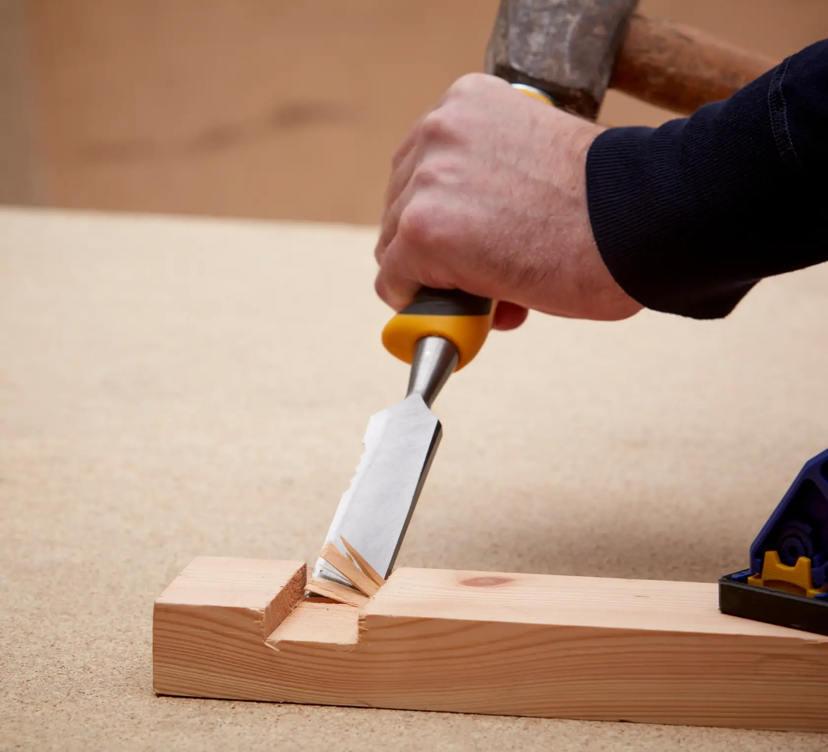 Hand using a DEWALT chisel and hammer to carve wood on a workbench.