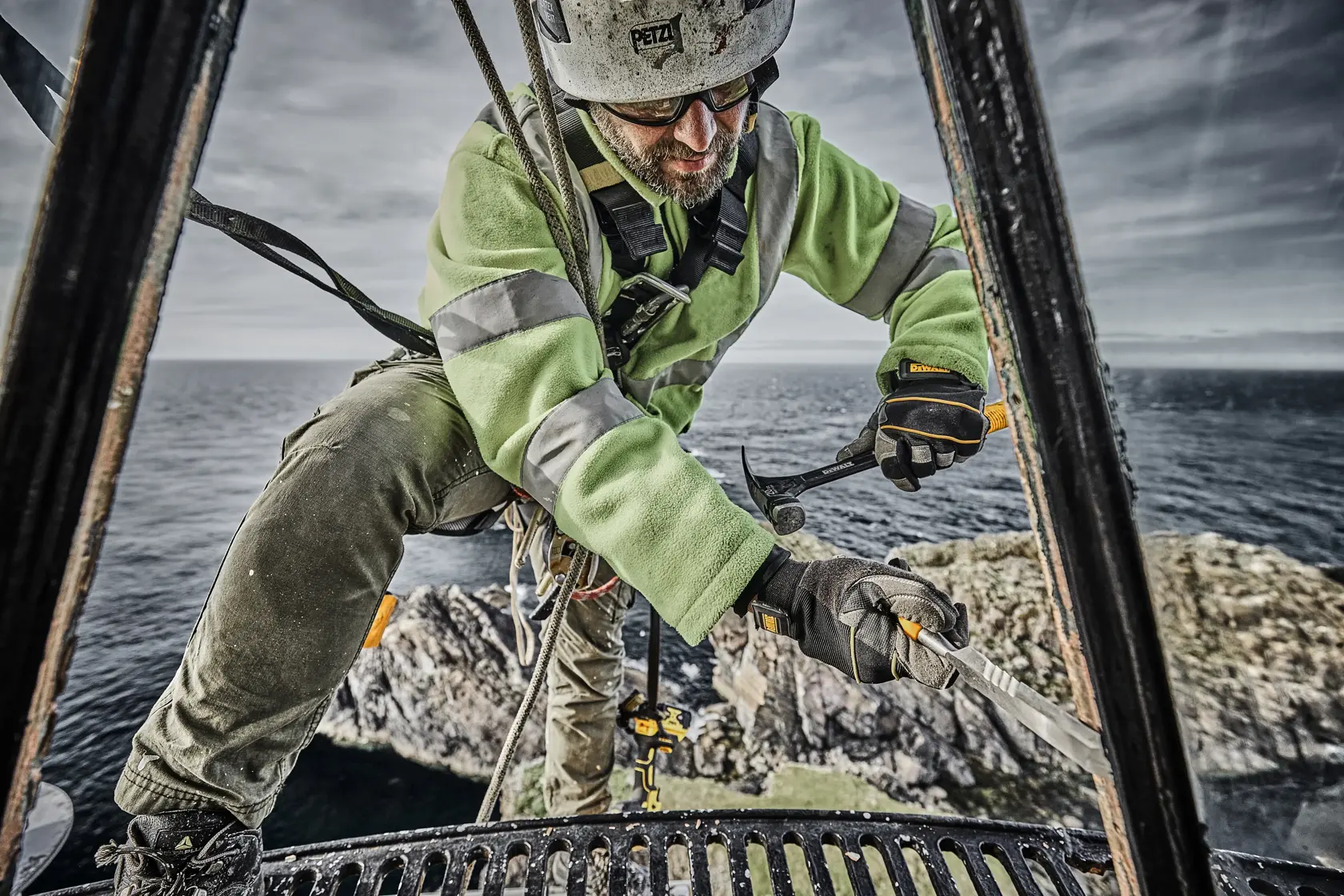 Worker in safety gear using a DEWALT tool outdoors near the sea, standing on metal scaffolding.