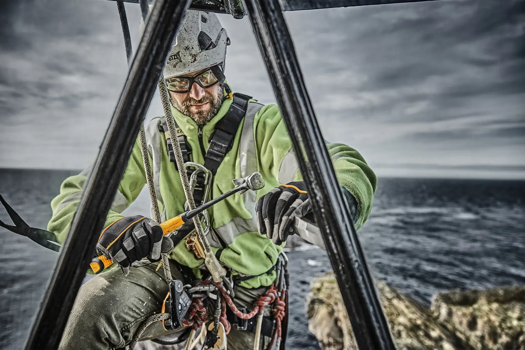 Worker with safety harness using a DEWALT tool on a structure outdoors near the sea.