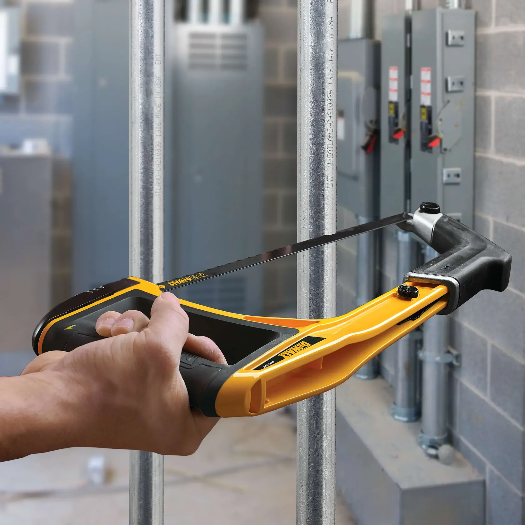 Hand holding a DEWALT hacksaw cutting a metal pipe in an industrial setting with electrical panels nearby.