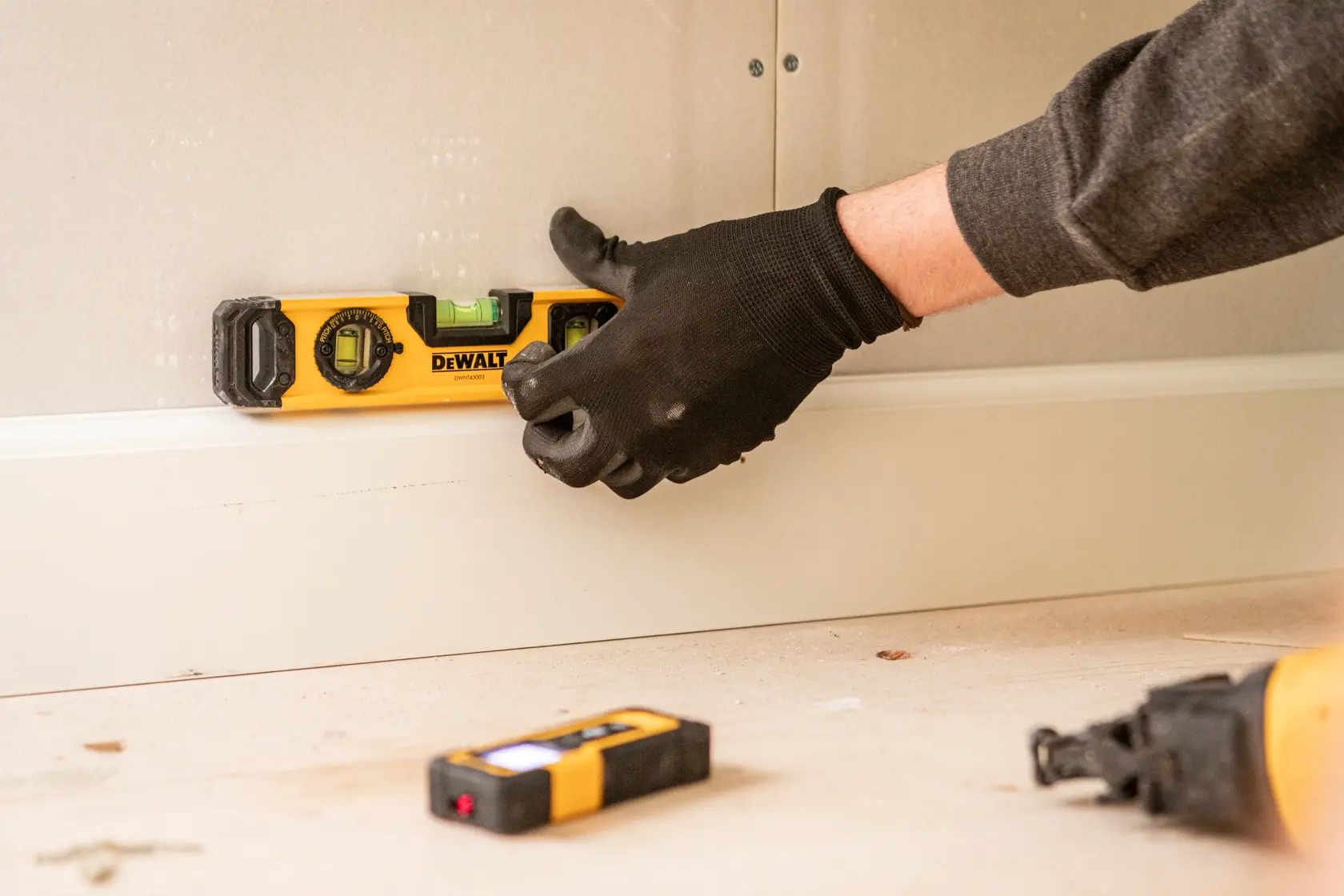 Person using DEWALT spirit level on wall trim, wearing black gloves. Other tools are visible on the floor.