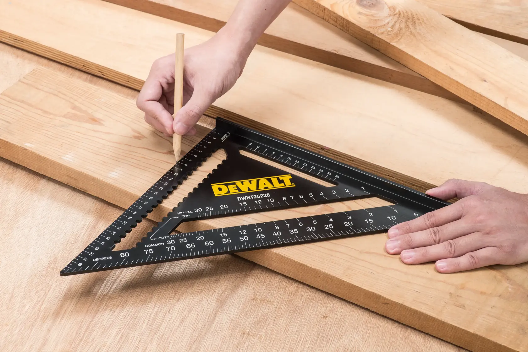 A DEWALT DWHT25228-0 photography square tool is placed on wooden boards. One hand holds the tool in position, while the other hand uses a pencil to mark measurements on the wood.