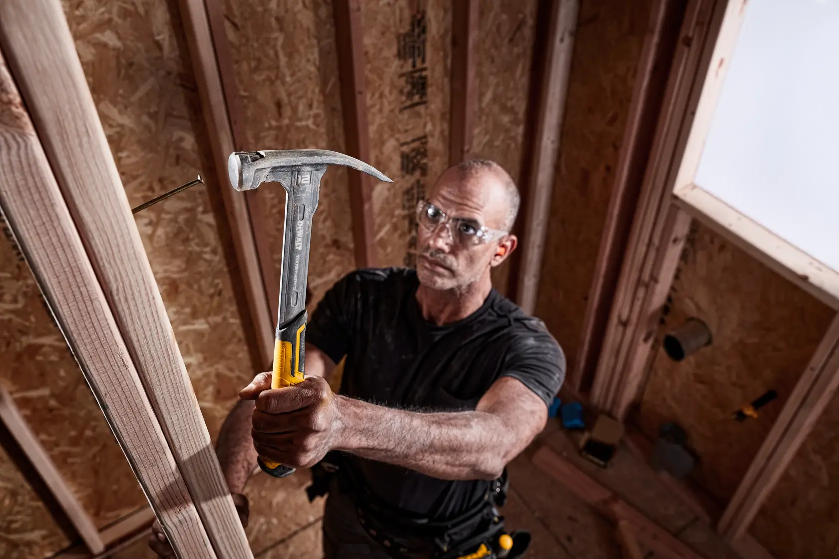 Una persona sta usando un martello DEWALT (modello DWHT51135-0) per piantare un chiodo in una struttura di legno all'interno di una stanza parzialmente costruita. Il martello ha un manico giallo e metallico.
