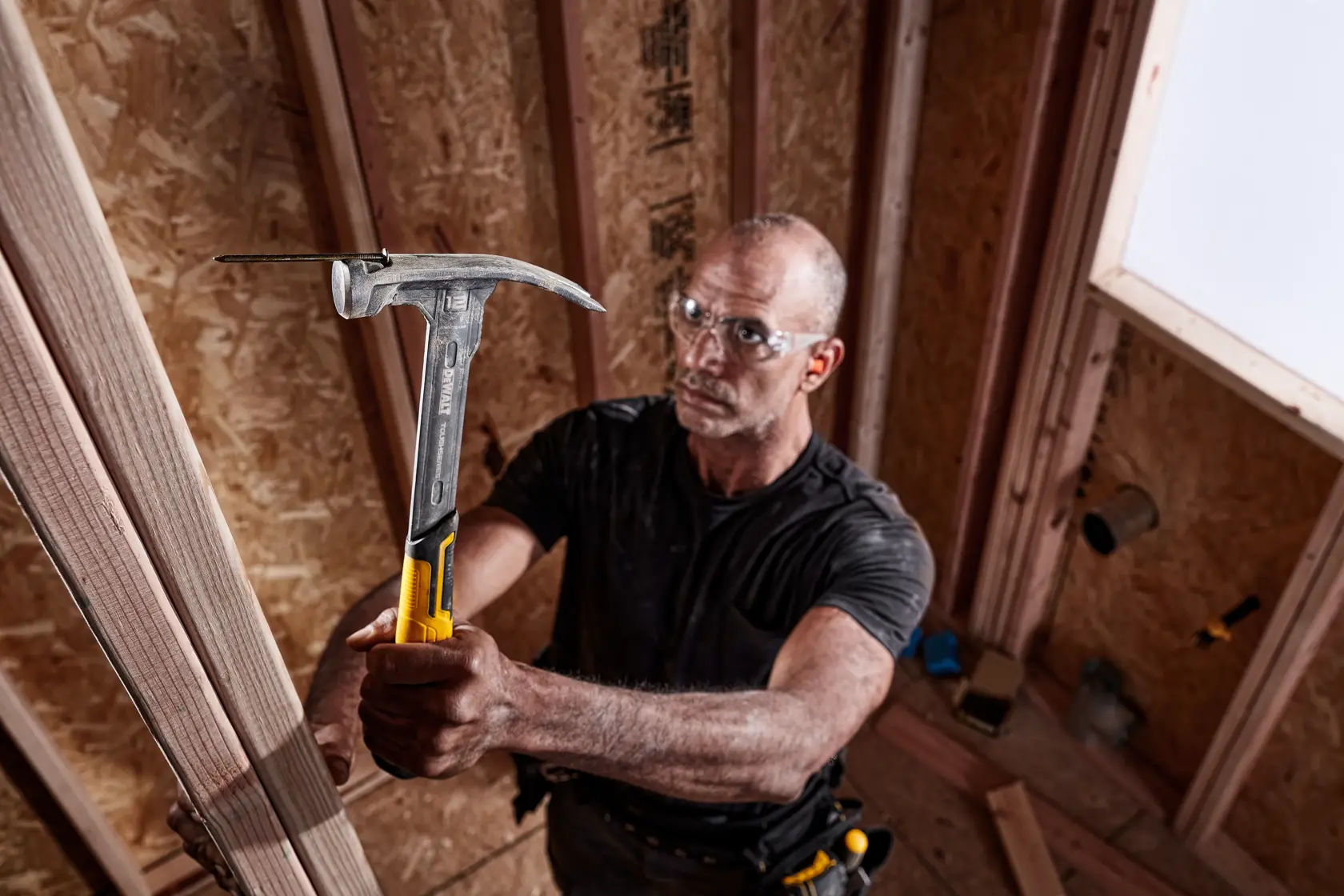 Una persona utilizza un martello DEWALT (DWHT51135-0) per lavorare su una struttura in legno all'interno di un cantiere. Il martello ha un manico giallo e nero e la persona indossa una cintura portautensili.