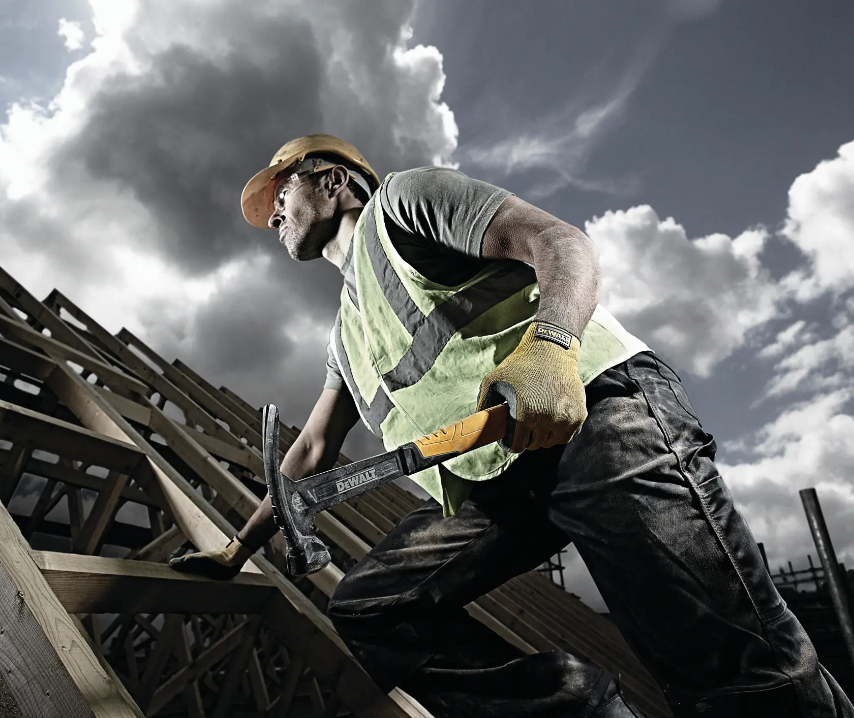 Operaio su un tetto con martello DEWALT, gilet, guanti e casco. Travi in legno e cielo nuvoloso.