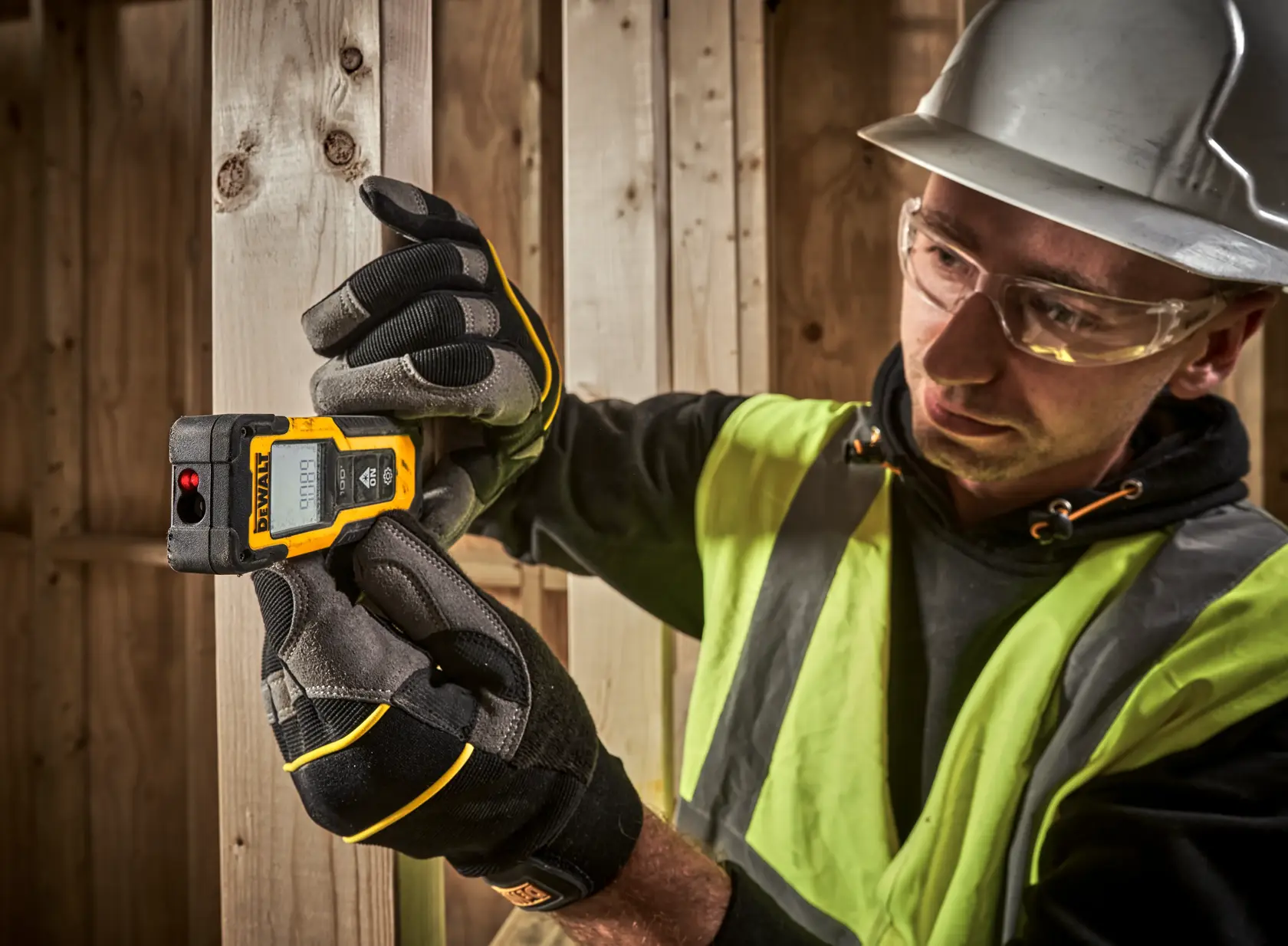 Worker uses DEWALT DWHT77100 laser distance measurer on wooden frame, wearing gloves and safety gear.