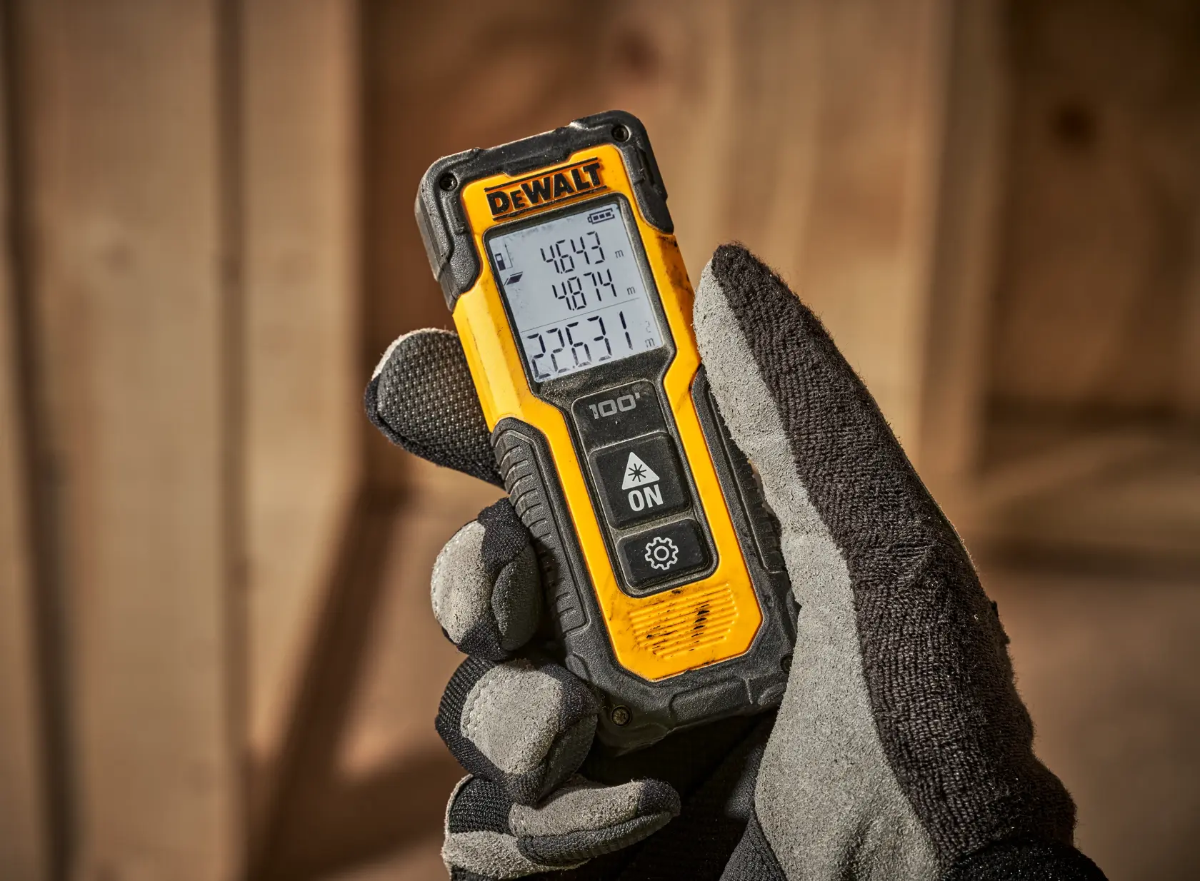 Gloved hand holding a DEWALT laser distance measuring tool with a digital display in a wooden workspace.