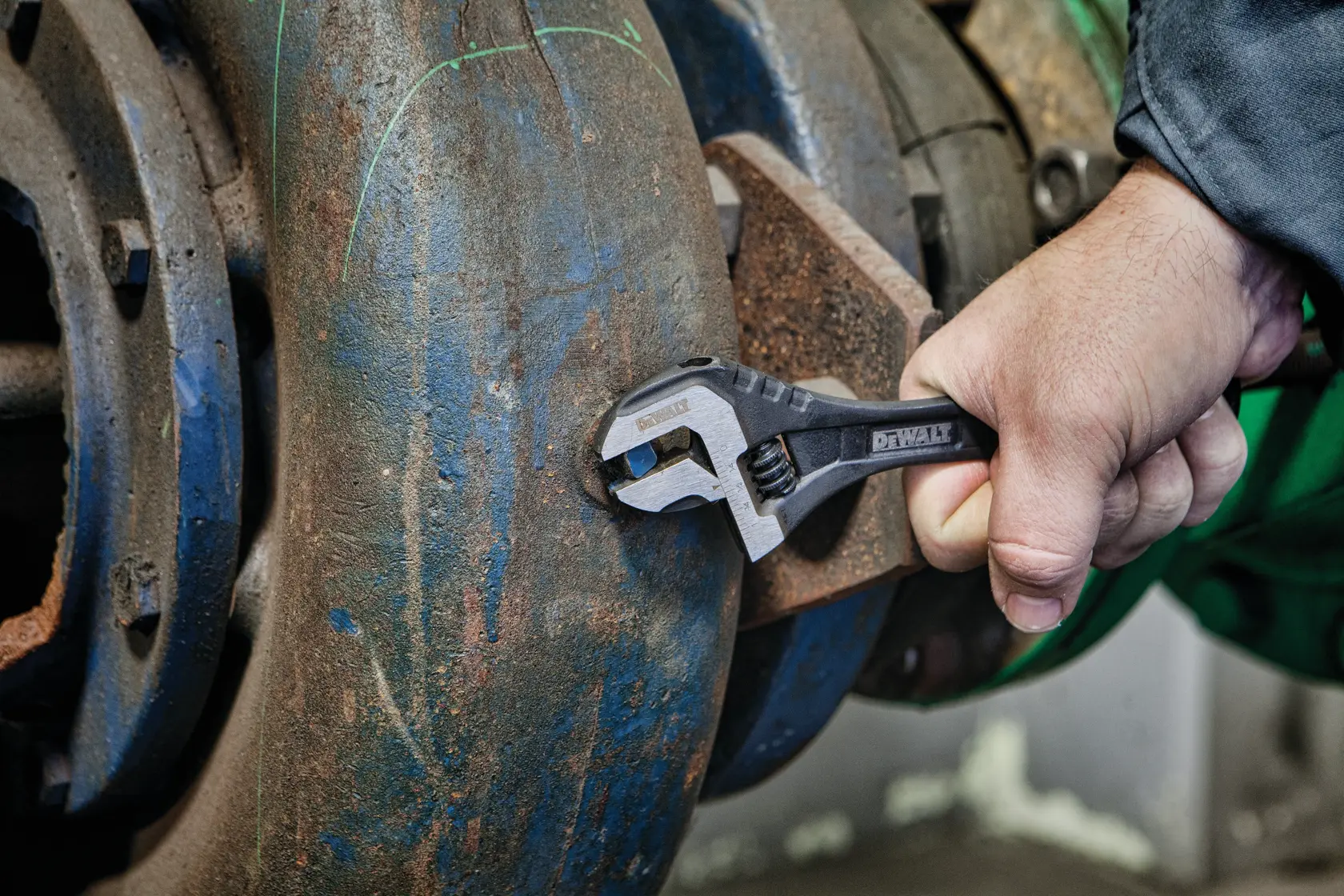 Hand using DEWALT adjustable wrench on a rusted pipe fitting in an industrial setting.