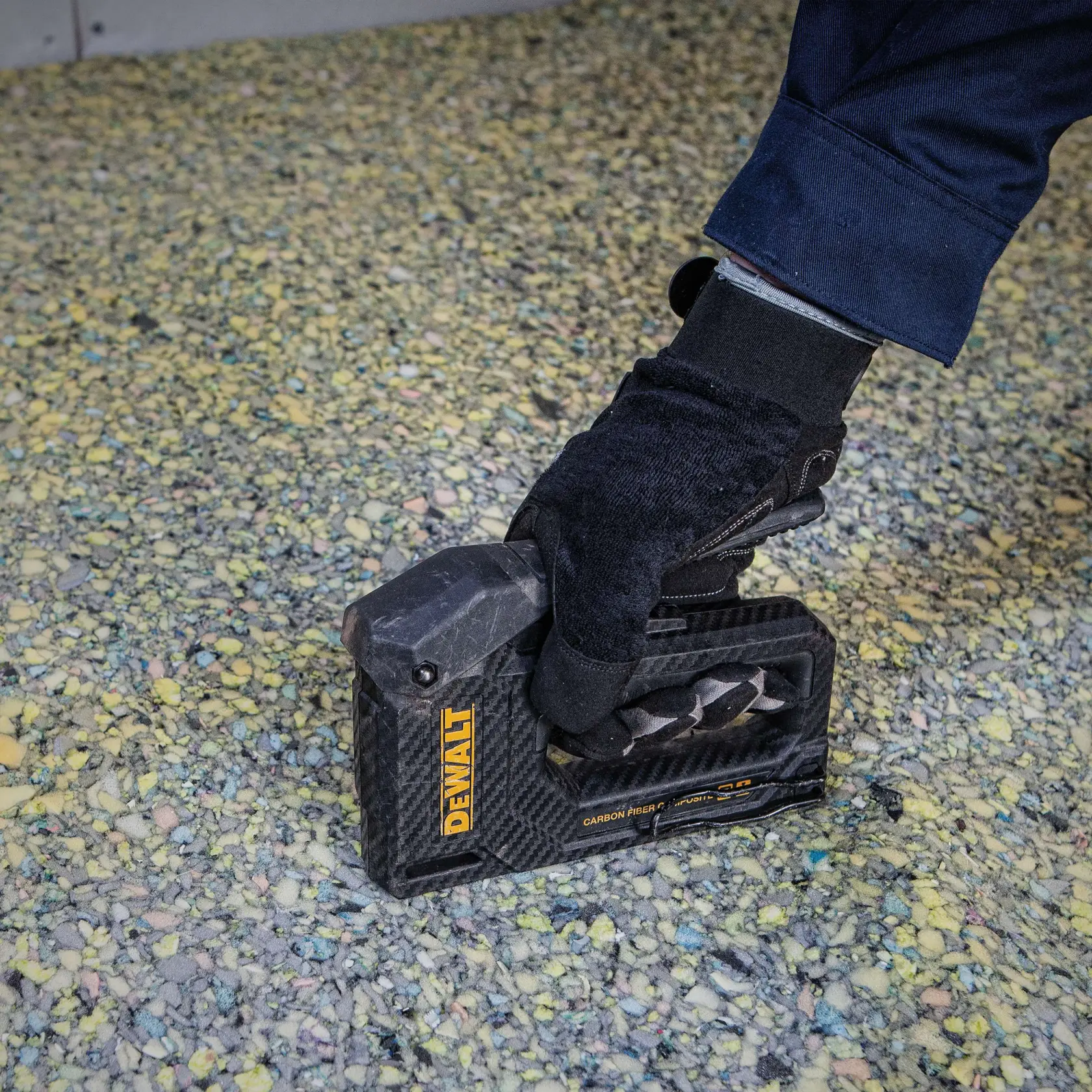 A gloved hand using a DEWALT carbon fiber staple gun to fasten material on a speckled surface.