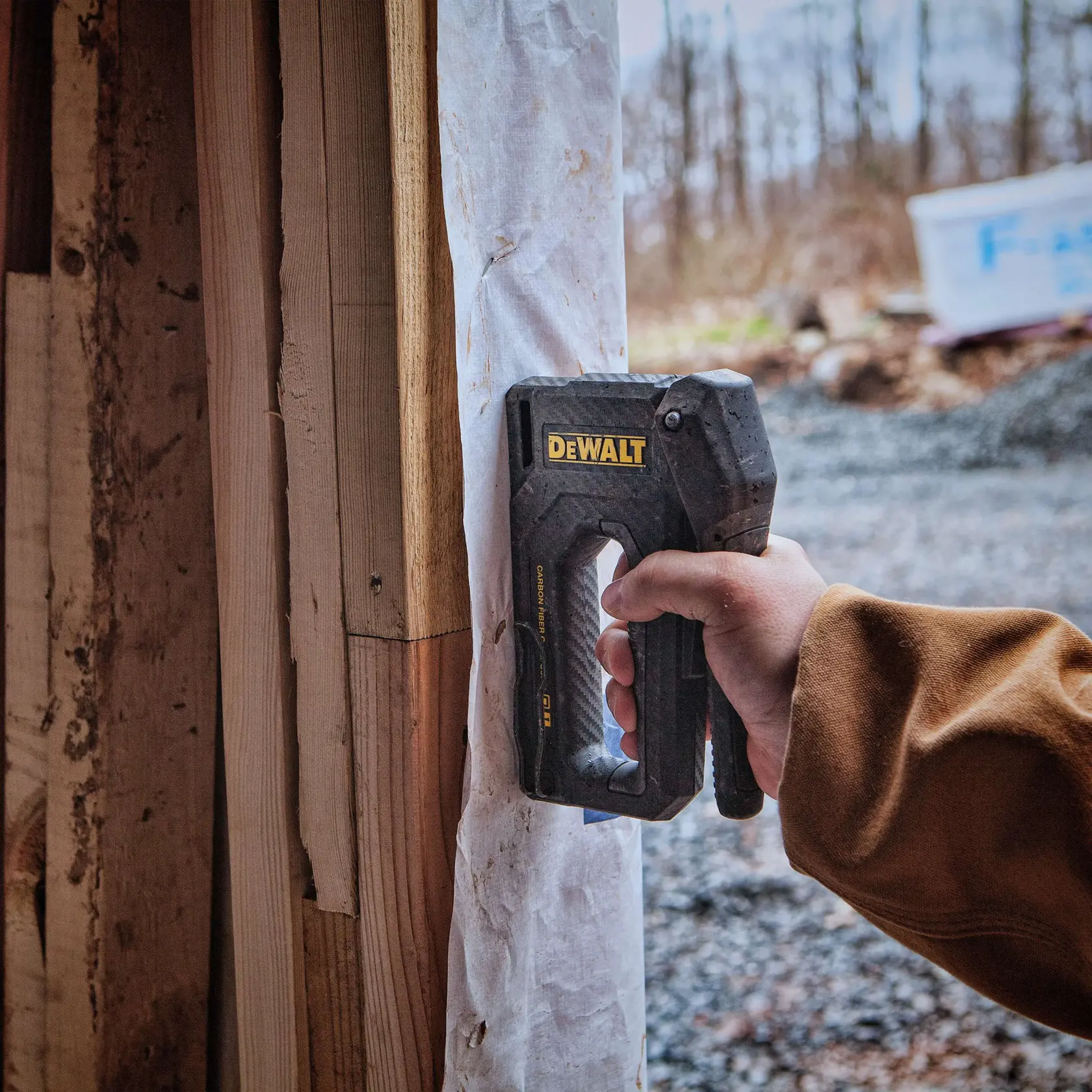 A person is using a DEWALT stapler, model DWHT80276, to secure a sheet of material to wooden framing at a construction site.