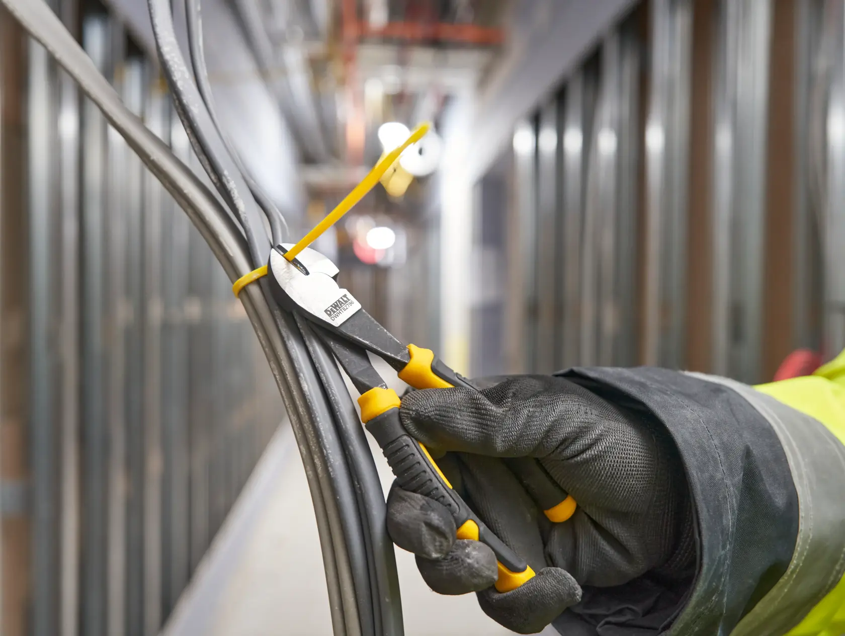 Gloved hand uses DEWALT pliers to cut a zip tie securing several bundled electrical cables in an industrial setting.