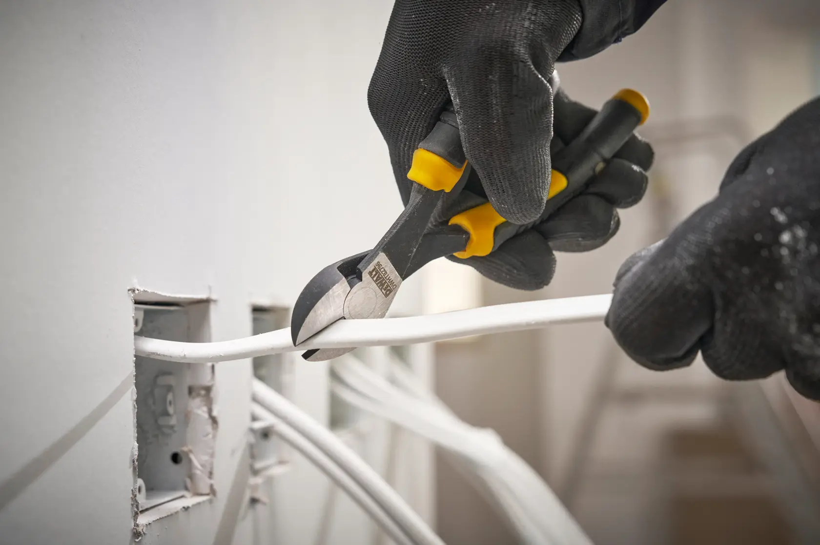 Gloved hands use DEWALT pliers to cut an electrical wire near a wall outlet with multiple cables.