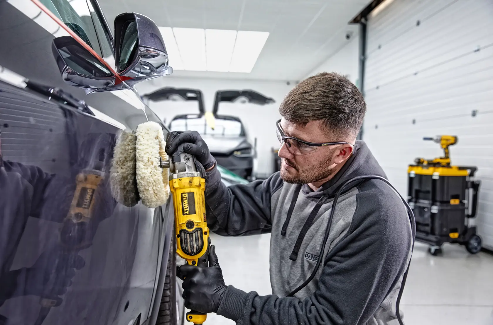 Person using a DEWALT DWP849X polisher on a car in a garage setting. Tool cart visible in background.