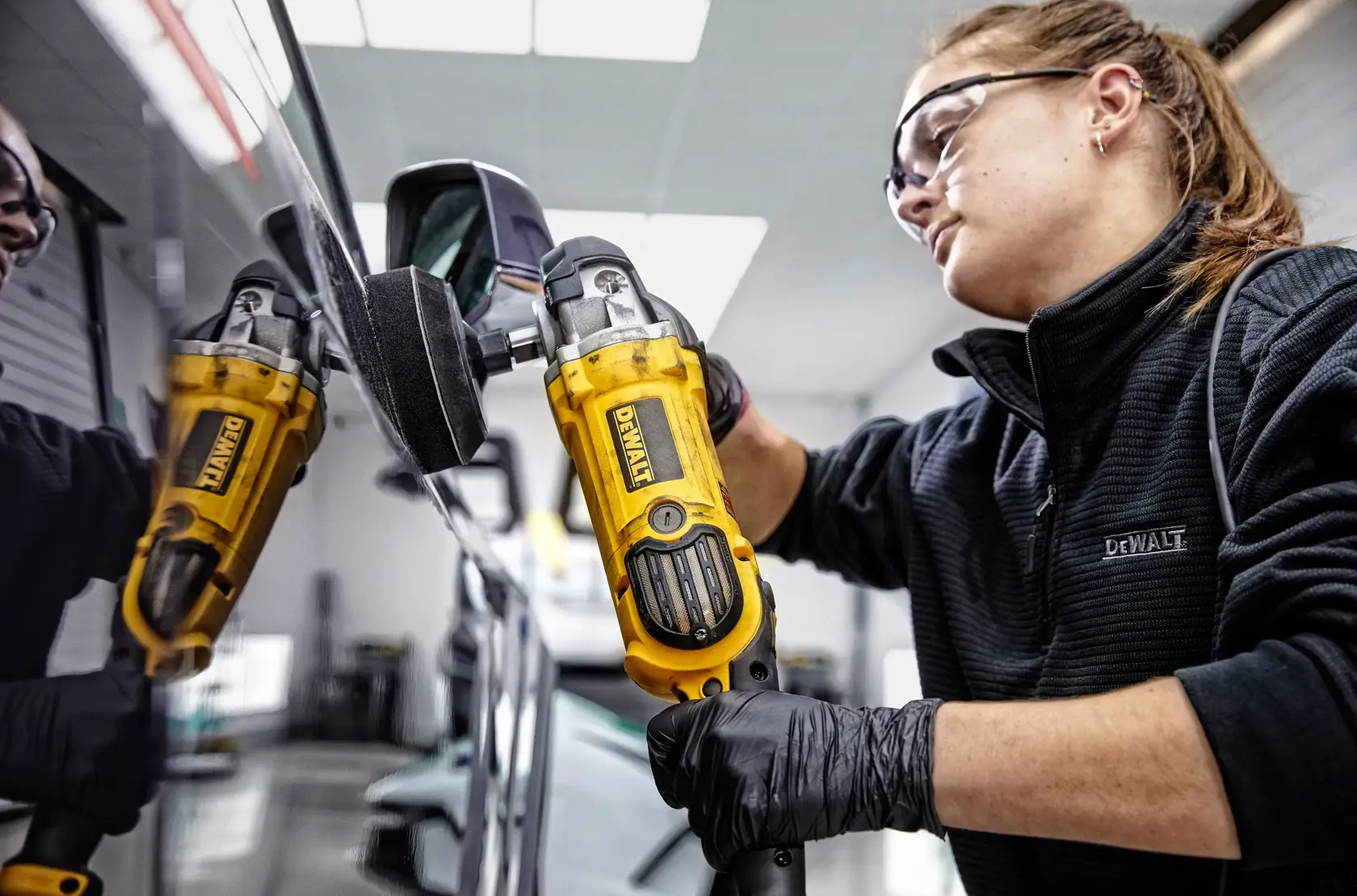 Person uses a yellow DEWALT polisher, model DWP849X, to polish a car side mirror indoors. Face is blurred.