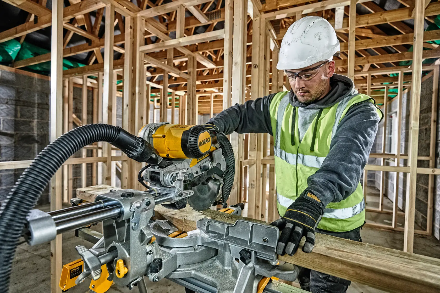 Worker uses a DEWALT miter saw to cut wood at a construction site, wearing safety gear and gloves.