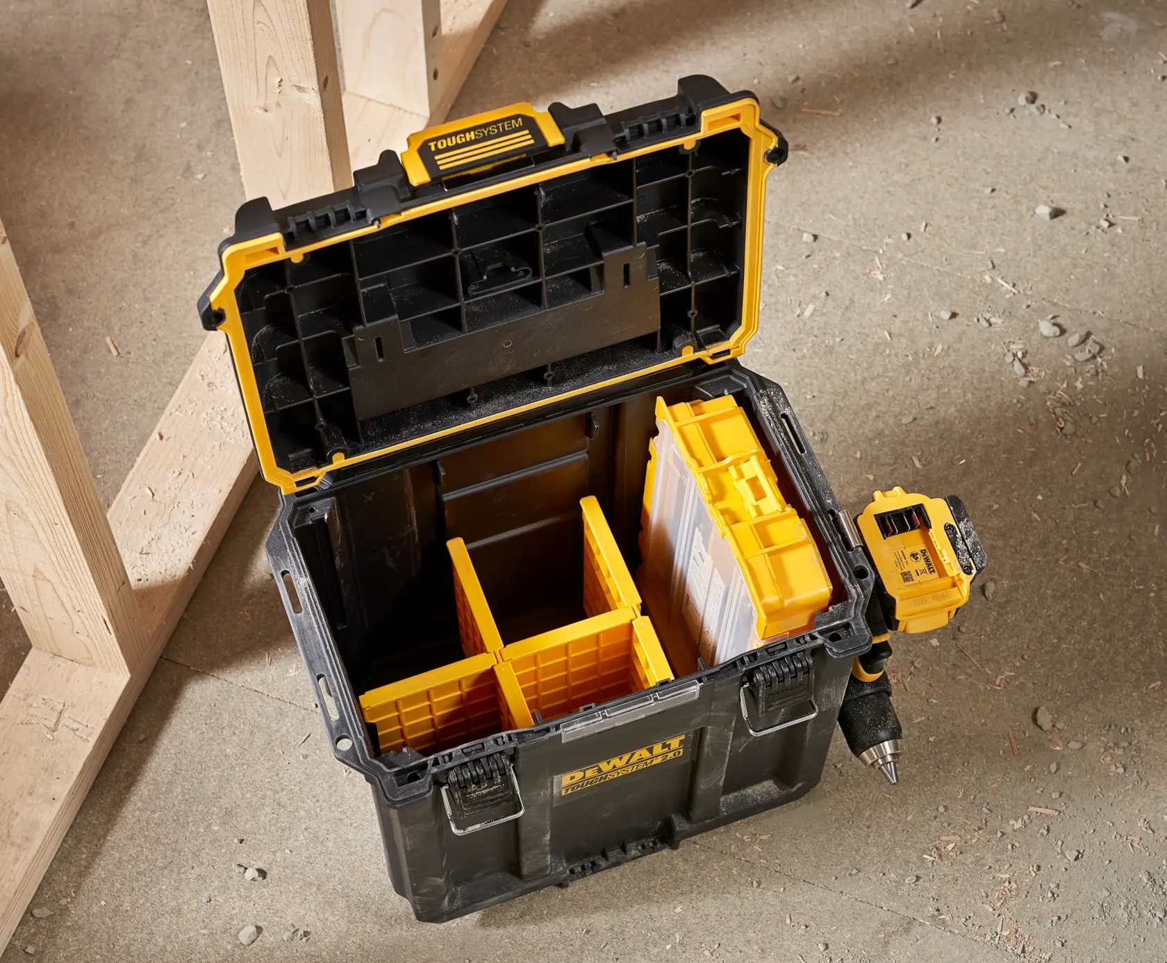 Åpen DEWALT ToughSystem oppbevaringsboks med gule rom og en drill festet på siden, plassert på et betonggulv ved siden av en treramme.
