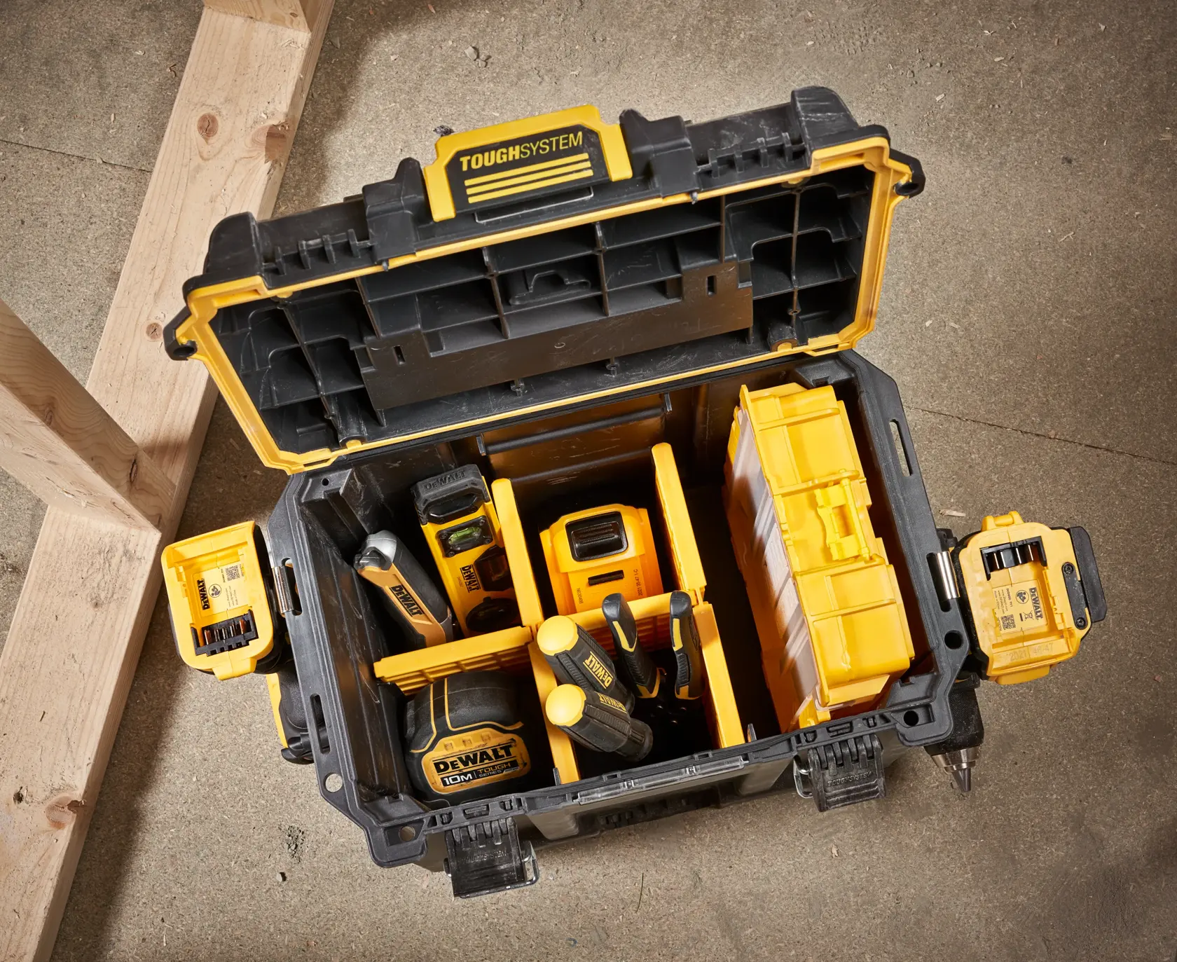 Åpen DEWALT ToughSystem verktøykasse som inneholder ulike gule og svarte verktøy og tilbehør, plassert på et betonggulv ved siden av trebjelker.