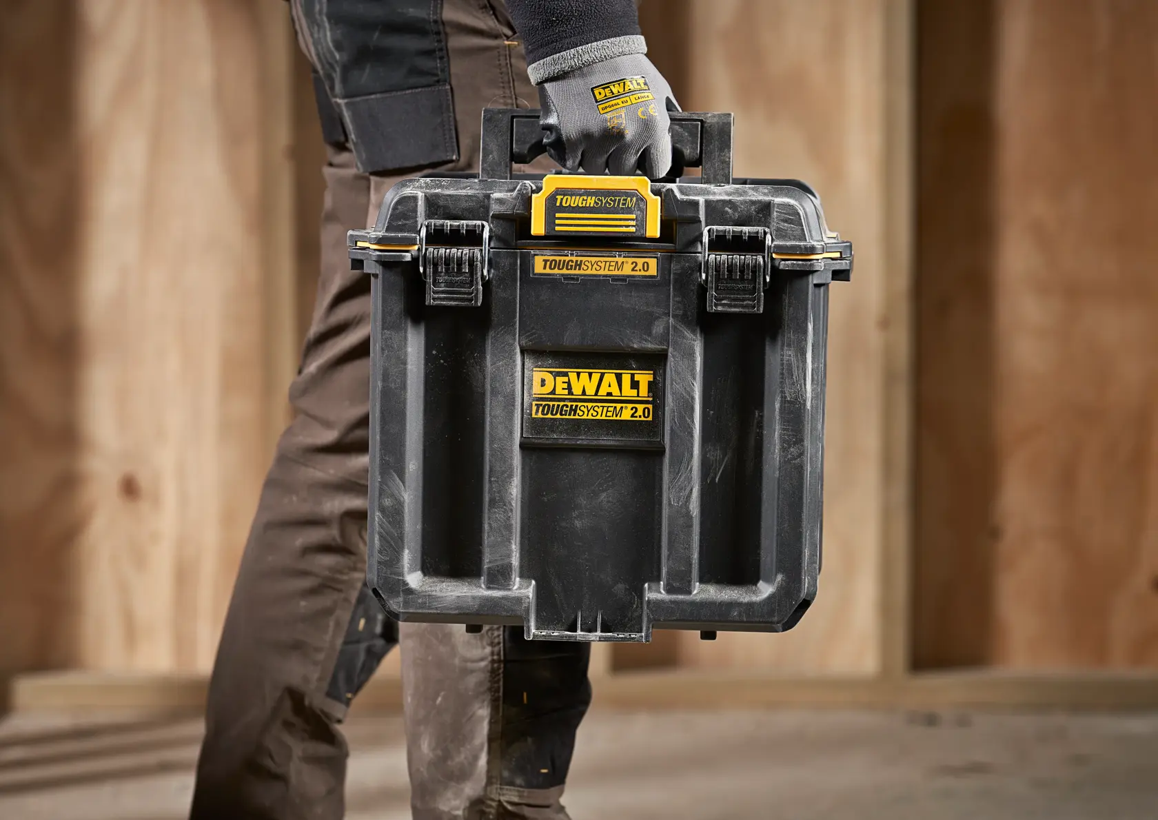 En person med arbeidshansker bærer en svart DEWALT TOUGHSYSTEM 2.0 oppbevaringsboks med gule etiketter, designet for verktøyorganisering og transport, i en byggeplass med trevegger.