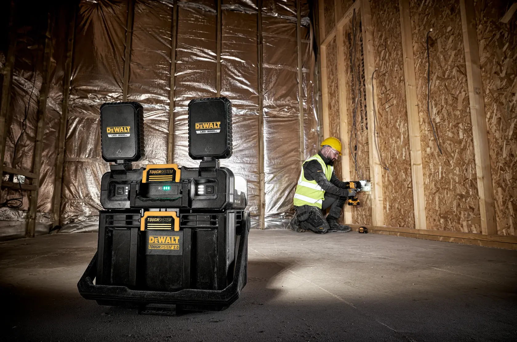 En DEWALT ToughSystem arbeidsradioset står på gulvet på en byggeplass med uferdige trevegger og isolasjon. I bakgrunnen jobber en arbeider med gul hjelm og refleksvest.
