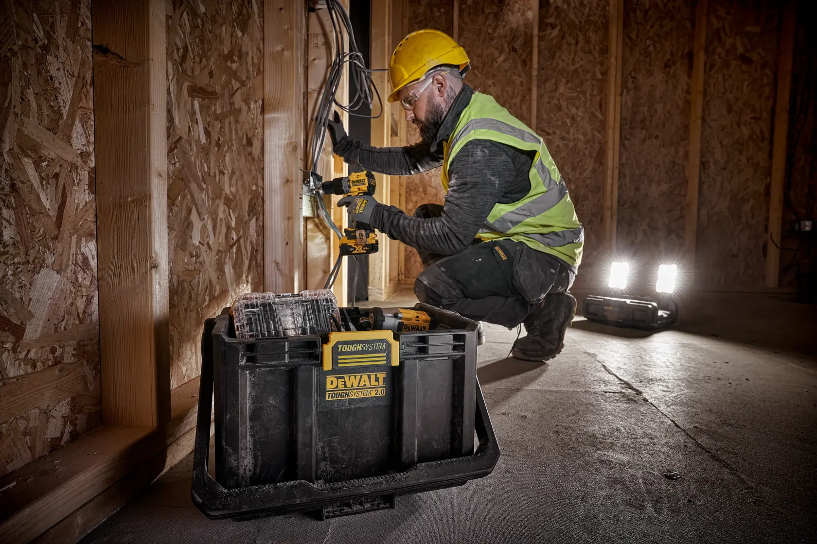 En person med gul hjelm og refleksvest bruker et DEWALT elektrisk verktøy for å arbeide med kabler på en trekonstruksjonsarbeidsplass. En DEWALT TOUGHSYSTEM 2.0 verktøykasse står i forgrunnen, og portable arbeidslys lyser opp området.
