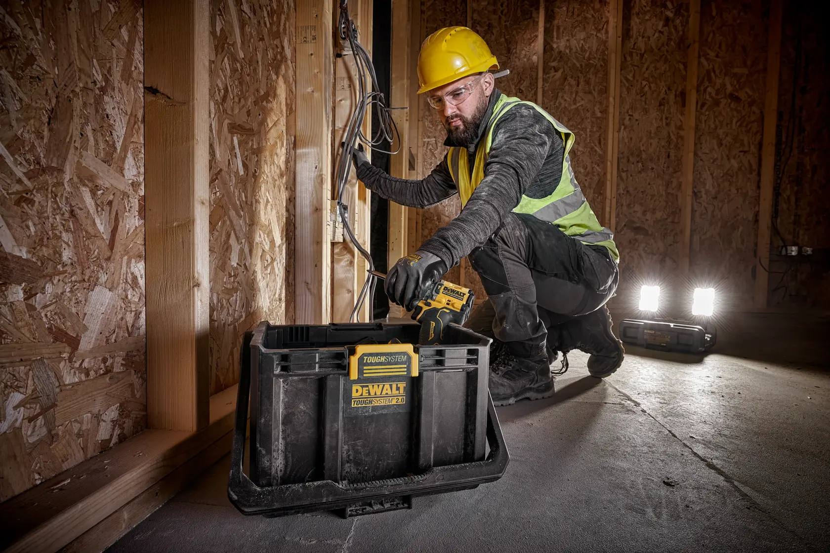 En person med gul hjelm og refleksvest arbeider med elektriske ledninger inne i en bygning med trekonstruksjon. Foran dem står en DEWALT ToughSystem oppbevaringsboks, og to portable arbeidslys er plassert på gulvet.