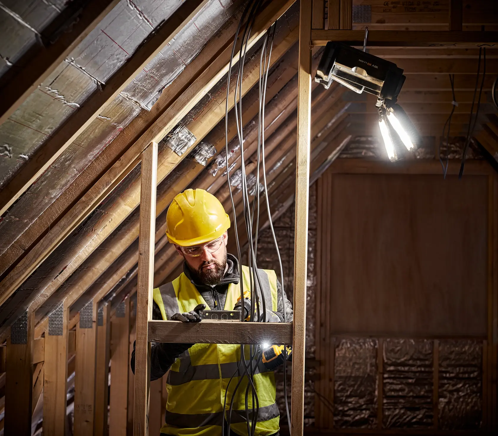 En bygningsarbeider med gul hjelm og refleksvest arbeider på et delvis ferdig loftsrom. Arbeideren håndterer verktøy og elektriske ledninger, mens en bærbar DEWALT arbeidslampe er festet over og lyser opp området.