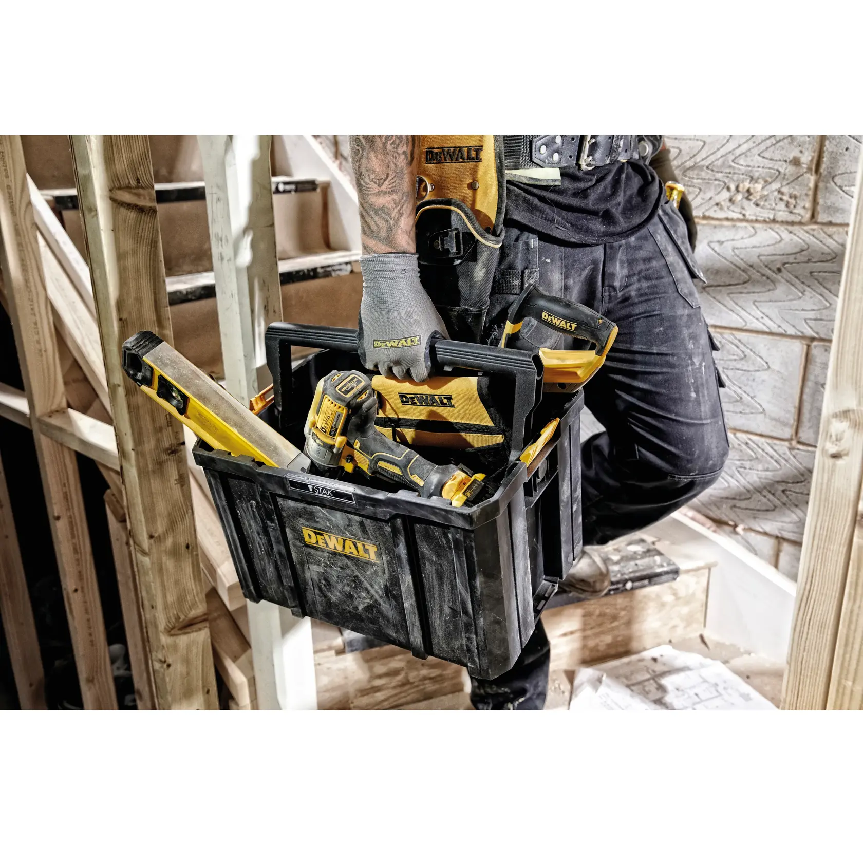 Person holding a DEWALT tool box with tools at a construction site. The box contains a drill and a level.