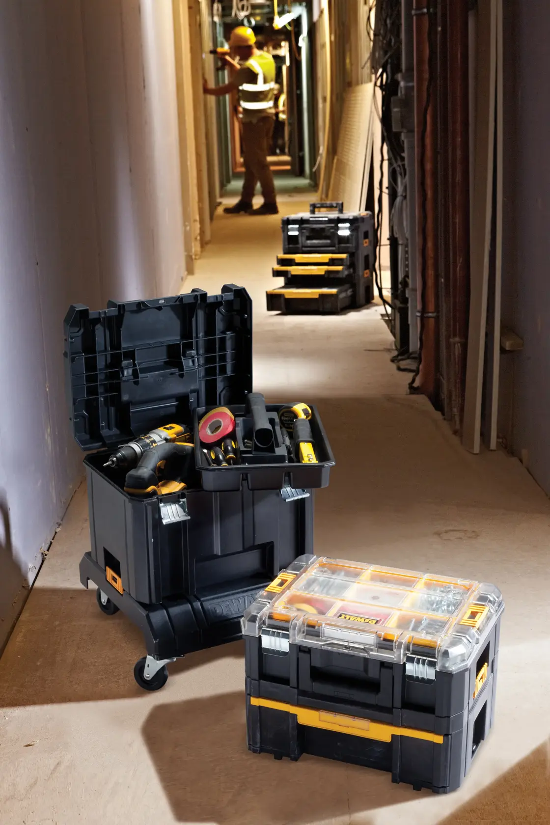 DEWALT DWST1-71229 tool storage boxes with tools, shown in a construction hallway with a worker in the background.