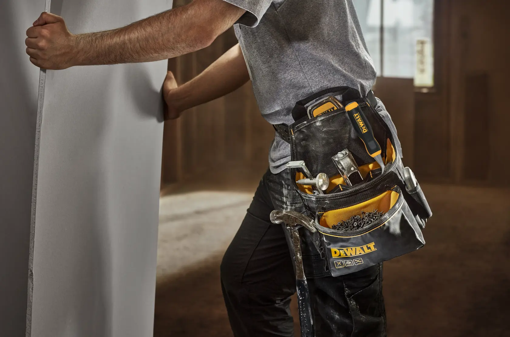 En person med DEWALT verktøybelte holder et stort panel. Verktøybeltet inneholder ulike håndverktøy, inkludert en hammer, skrutrekker og festemidler. Scenen er innendørs og antyder et bygge- eller installasjonsmiljø.
