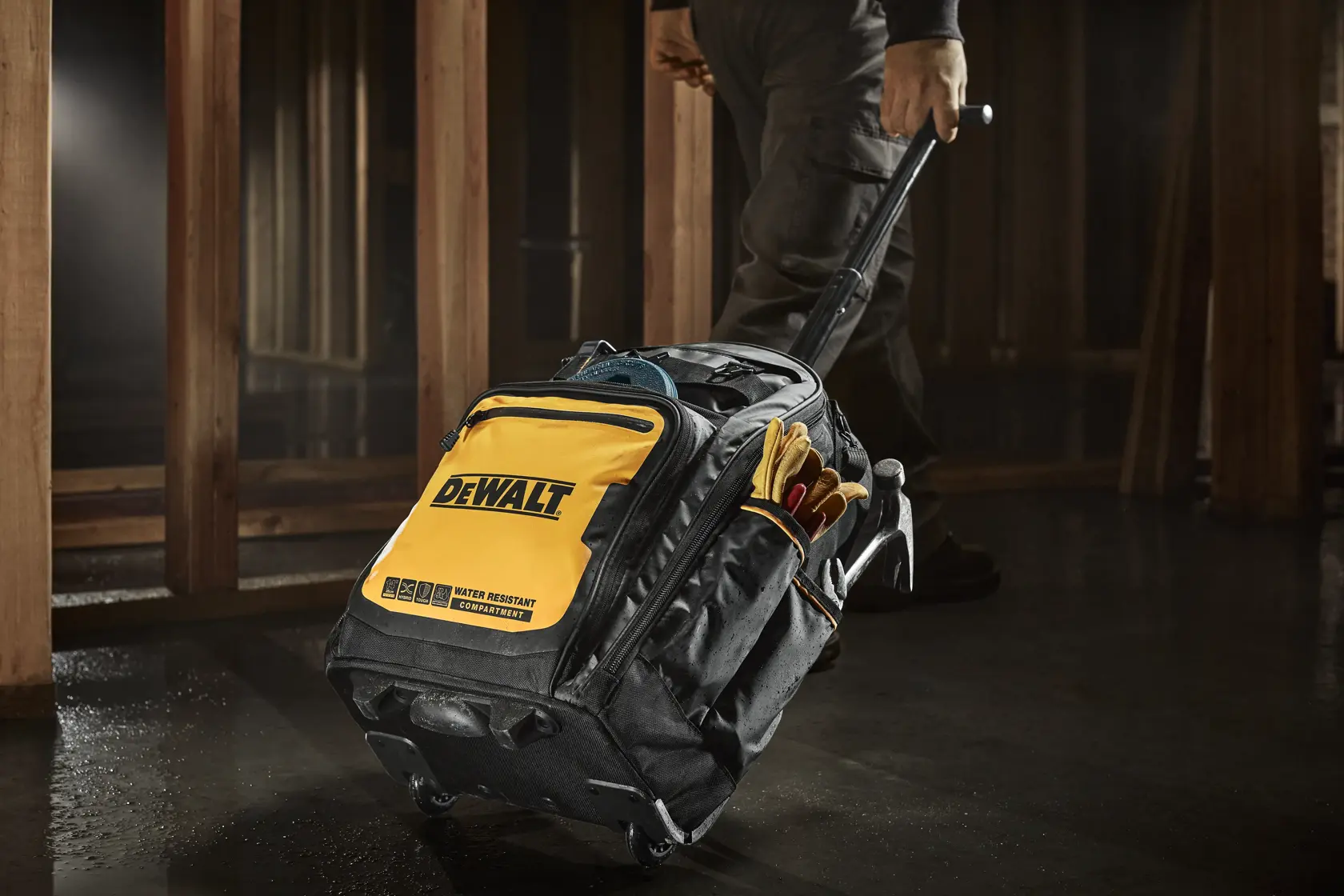 En svart og gul DEWALT verktøybag med hjul og uttrekkbart håndtak, trekkes av en person i et arbeidsmiljø. Bagen har ytterlommer med arbeidshansker og annet tilbehør til verktøy.