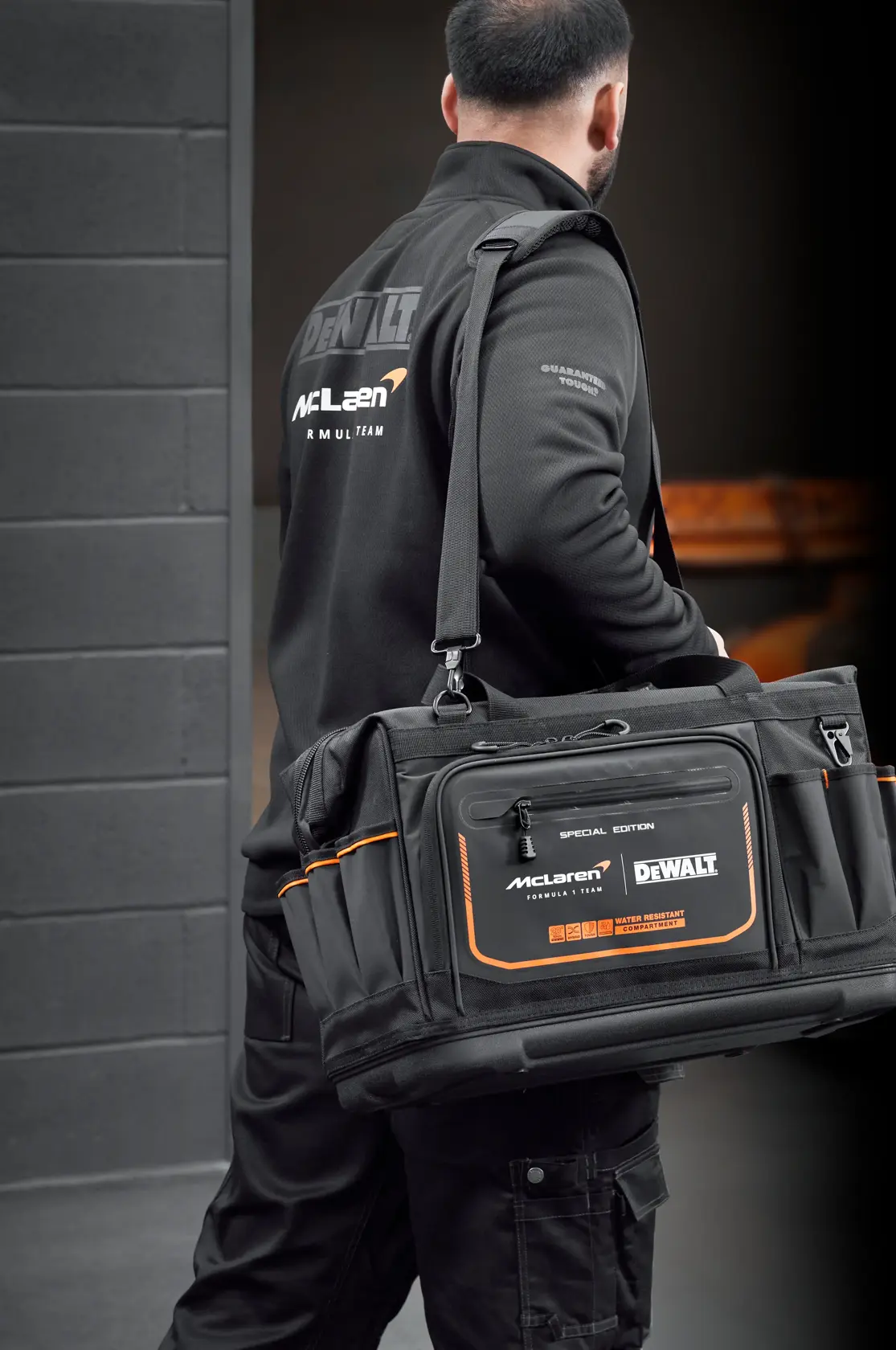Person carrying a black DEWALT McLaren tool bag with orange accents in an industrial setting.